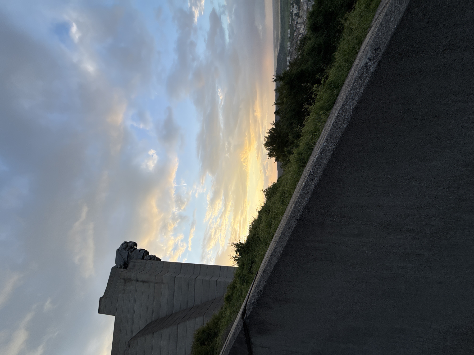 The image depicts the Founders of the Bulgarian State Monument in Shumen, Bulgaria. The monument is made of concrete and features a sculpted figure at its peak. The sky is filled with clouds illuminated by a sunset. Vegetation covers the slope behind a concrete wall in the foreground. A portion of the city of Shumen is visible in the distance.