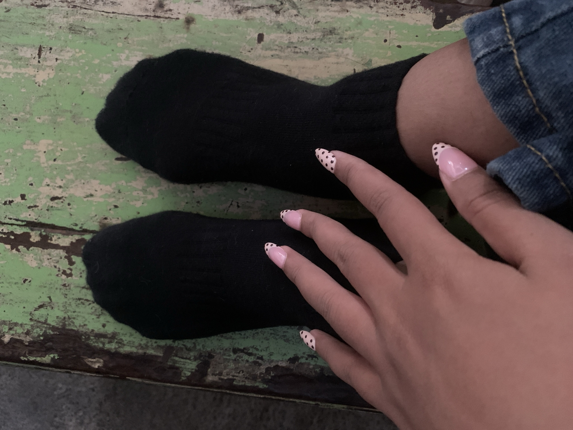 Two feet, clad in black socks, rest on a weathered, green wooden surface. The left foot is positioned slightly forward and to the left of the right foot. A person's hand, with light pink nails adorned with black polka dots, is visible in the foreground, resting on their leg. The denim of their jeans is also visible at the bottom right of the frame. The scene appears to be indoors, given the lighting and the context of resting feet. It's not possible to determine the specific location within Tamanbaru, Indonesia from the image.