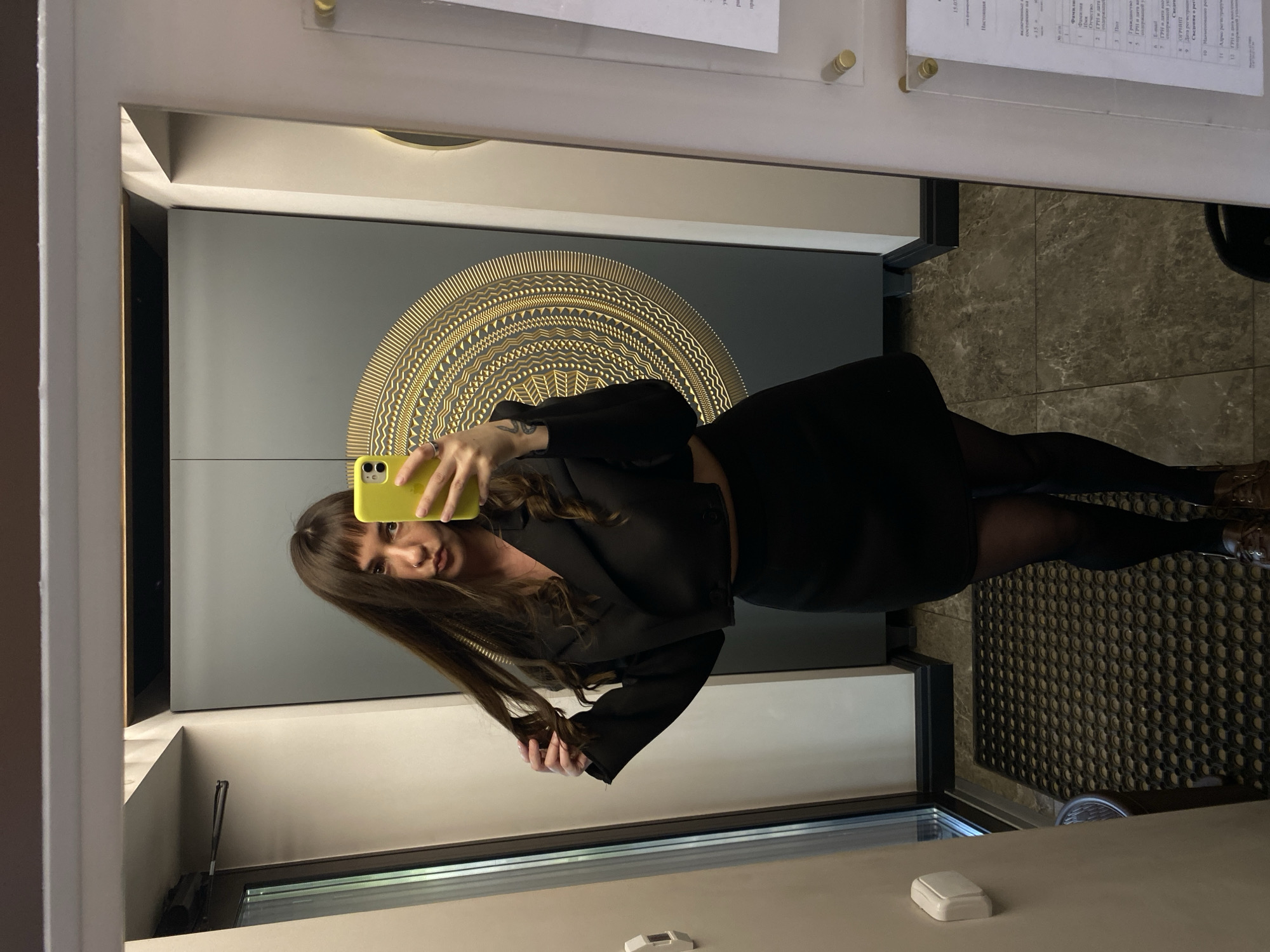 An interior mirror selfie taken in what appears to be an administrative office, business reception area, or public service center in Votkinsk, Russia. A young woman with long, straight dark brown hair and bangs is the central subject, holding a bright yellow smartphone to capture her reflection. She is making a slight pouting expression and uses her left hand to hold a section of her hair. She wears a black cropped blazer or jacket, a black mini skirt, sheer black tights, and brown lace-up shoes.
The background, visible in the mirror's reflection, reveals a modern and well-maintained interior. Directly behind the woman, on a large gray panel or double door, is a prominent, circular golden geometric design, reminiscent of a stylized sun or mandala. To the left of the reflection (viewer's right), a glass-paned door or window provides a glimpse of outdoor greenery, suggesting an entrance. Below this, a dark, textured floor mat is visible, covering part of the tiled floor. The floor throughout the area consists of polished dark gray or brown tiles, possibly marble or granite. To the right of the reflection (viewer's left), a white wall features a clear acrylic information display board, mounted with golden standoffs. Several sheets of paper with Cyrillic text are visible on this board, containing fields such as "Фамилия" (Last Name), "Имя" (First Name), "Отчество" (Patronymic), "ГРН и дата" (GRN and date), "ИНН" (Taxpayer ID), "Гражданство" (Citizenship), "E-mail," and "Сведения о регистрации" (Information about registration), indicating its use for official or business-related communication. The lighting appears to be artificial, casting soft illumination on the scene.