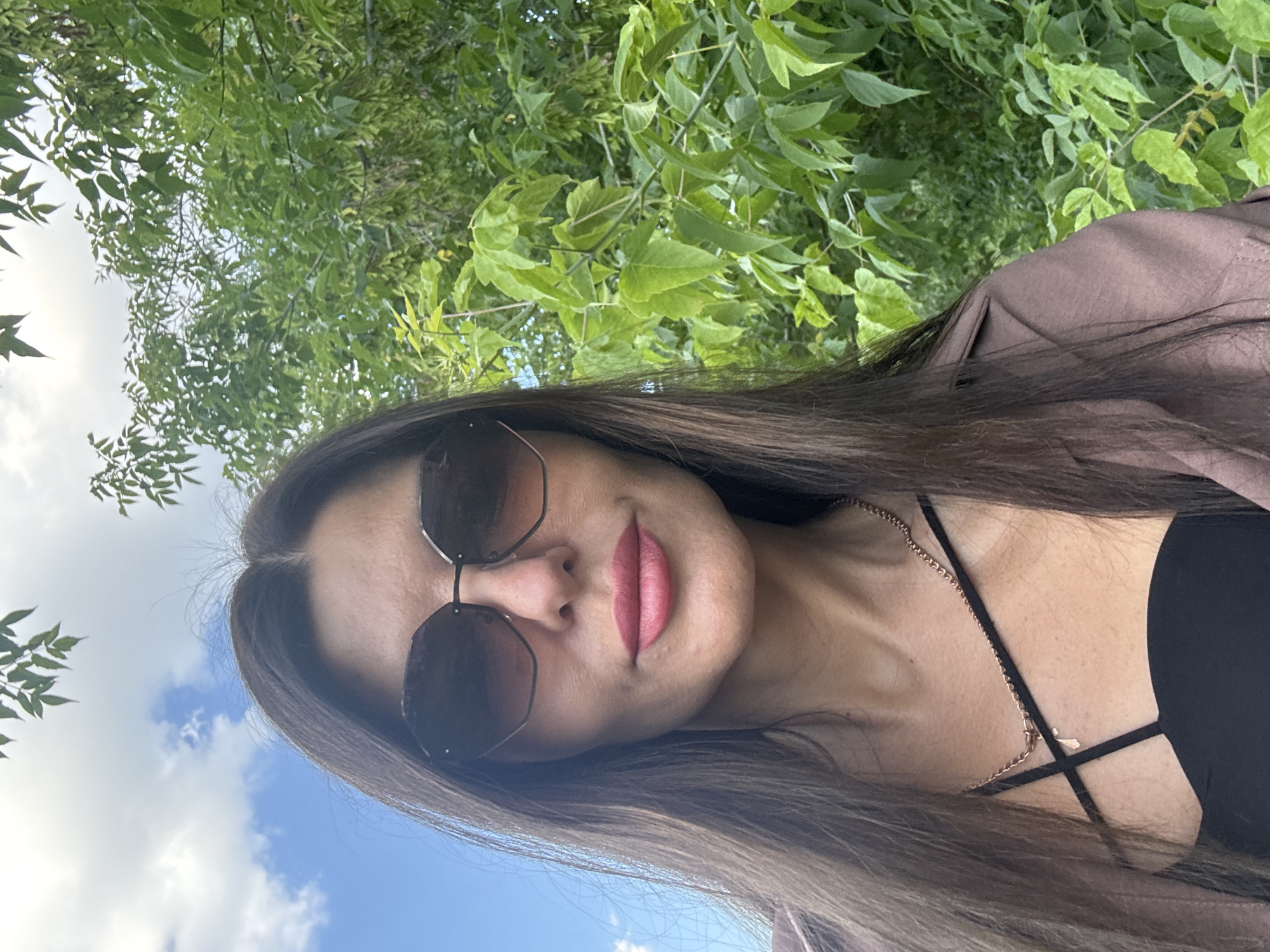 The media file shows a selfie of a woman with long, dark hair and sunglasses, taken outdoors. She is wearing a brown jacket over a black top, and a delicate gold necklace is visible around her neck. Her lips are painted a rosy pink, and she has a subtle smile. The background is filled with lush green foliage, suggesting a natural setting, possibly a park or garden, with a bright blue sky and scattered clouds visible at the bottom. The lighting indicates it is daytime, likely a sunny afternoon. The orientation of the image is slightly tilted. No text is visible in the image.