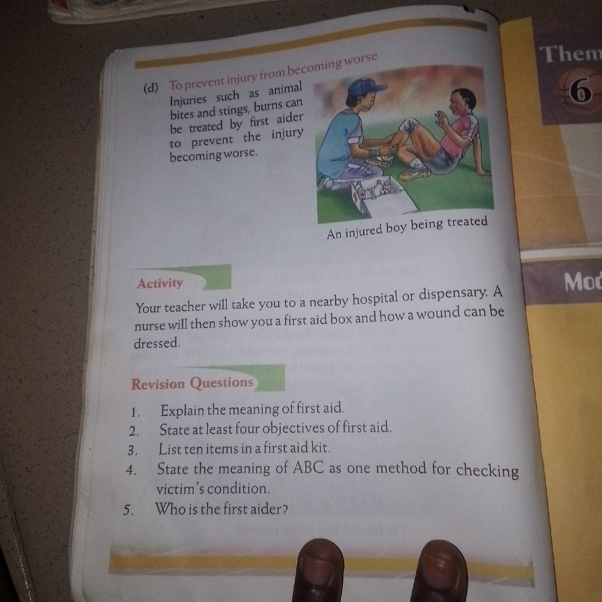 A book is open to a page with text and an illustration. The text on the left side of the page is divided into sections titled "Activity" and "Revision Questions". The "Activity" section describes a teacher taking students to a hospital or dispensary and showing them a first aid box. The "Revision Questions" section lists five questions related to first aid. The illustration on the right side depicts a boy in a blue shirt and cap attending to an injured boy in a pink shirt who is sitting on the ground. There is a first aid box open between them, containing various medical supplies. A caption below the illustration reads "An injured boy being treated". The right edge of the page shows text with "Theme" and "6" on a vertical banner, and below that, "Mod". The overall scene appears to be a page from an educational book, likely for children, focusing on first aid.