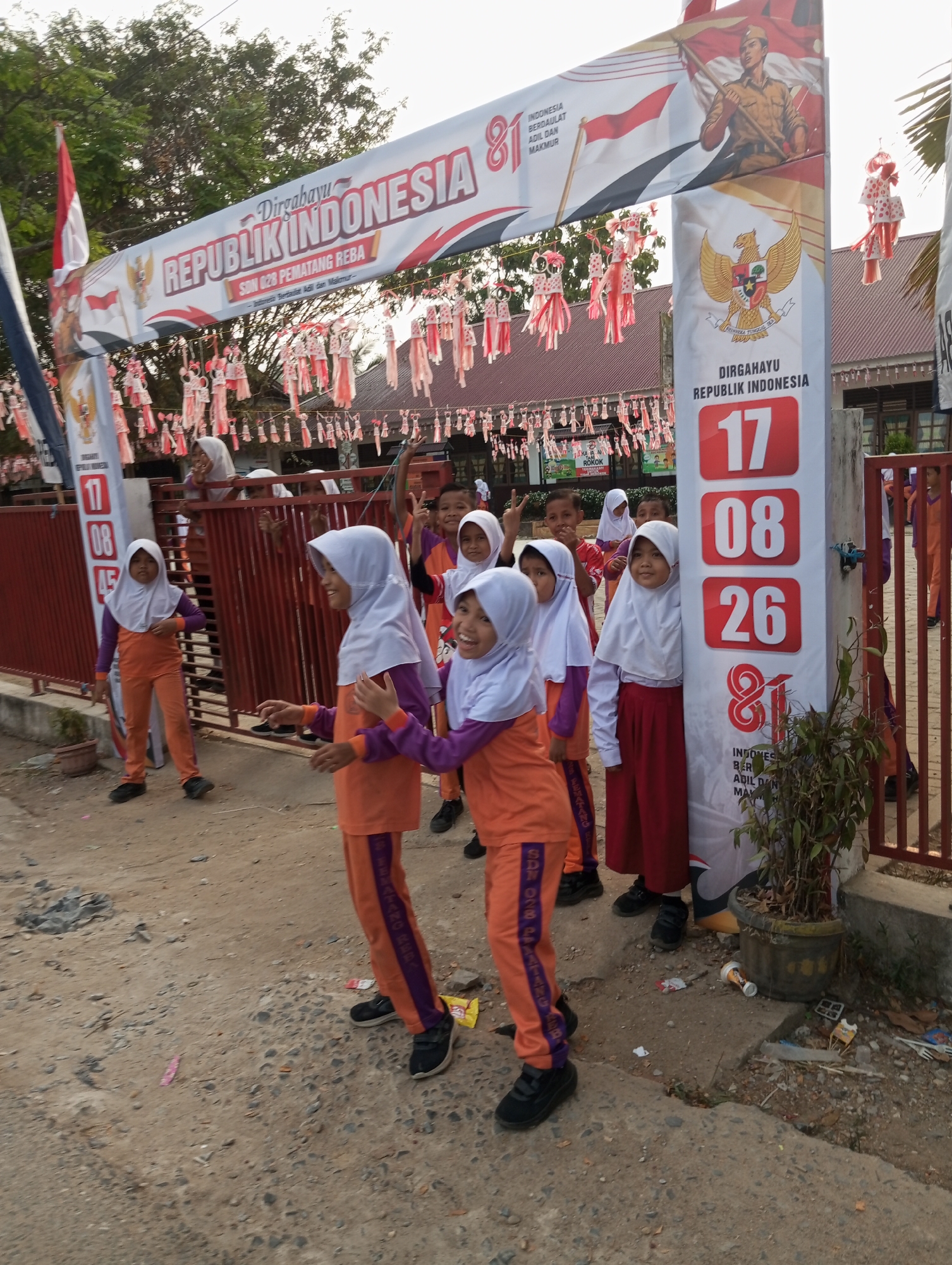 The image depicts an outdoor scene in Pematang Reba, Indonesia, likely near an educational institution given the presence of children. A large banner with Indonesian national symbols and text, including "Dirgahayu Republik Indonesia," is prominently displayed, possibly for a celebration. Children, predominantly girls wearing orange uniforms with purple accents and headscarves, are gathered around the banner and a red metal fence. Some children appear to be posing or looking towards the camera, while others are engaged in playful interactions. The ground is a mix of dirt and gravel, with some scattered debris. The background shows buildings with red roofs, trees, and other decorative elements hanging from the banner. A potted plant is visible near a concrete structure. The overall atmosphere suggests a community event or school activity.