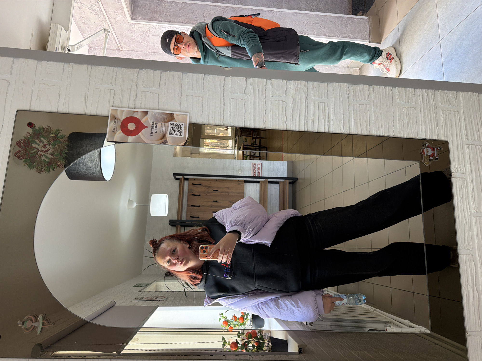 In an indoor setting in Noviy, Russia, a woman takes a mirror selfie while a man stands in the background. The primary subject, a woman with reddish-brown hair in two buns, is reflected in a large, rectangular mirror. She wears a black hoodie, a light purple puffer jacket, and black pants. She holds a smartphone to take the picture, pouting slightly, and carries a small plastic water bottle in her other hand, which rests on a low shelf.
The mirror's reflection shows a wooden cabinet and a white square light fixture behind her. Christmas decorations, including a wreath and a small Santa Claus figure, adorn the mirror's frame. On the mirror, a white sign displays Russian text: "Яндекс Карты" (Yandex Maps) and "Оставьте отзыв" (Leave a review), accompanied by QR codes. Below this, a partial, inverted reflection of another sign reads "САЛЕЙРУЖ" and "ОСЛЮДОННЕ", likely parts of "СДЕЛАЙТЕ СЕЛФИ" (Take a selfie) and "ПОЛУЧИТЕ ПОДАРОК" (Get a gift).
In the background, standing in what appears to be a doorway, a man is visible from the waist up. He wears a green tracksuit, a black vest over an orange backpack, a black cap, and orange-tinted sunglasses. He has a watch on his left wrist. The surrounding walls feature a textured, white brick-like pattern, and the floor in the background is tiled with light blue/grey and beige squares. The lighting suggests it is daytime.