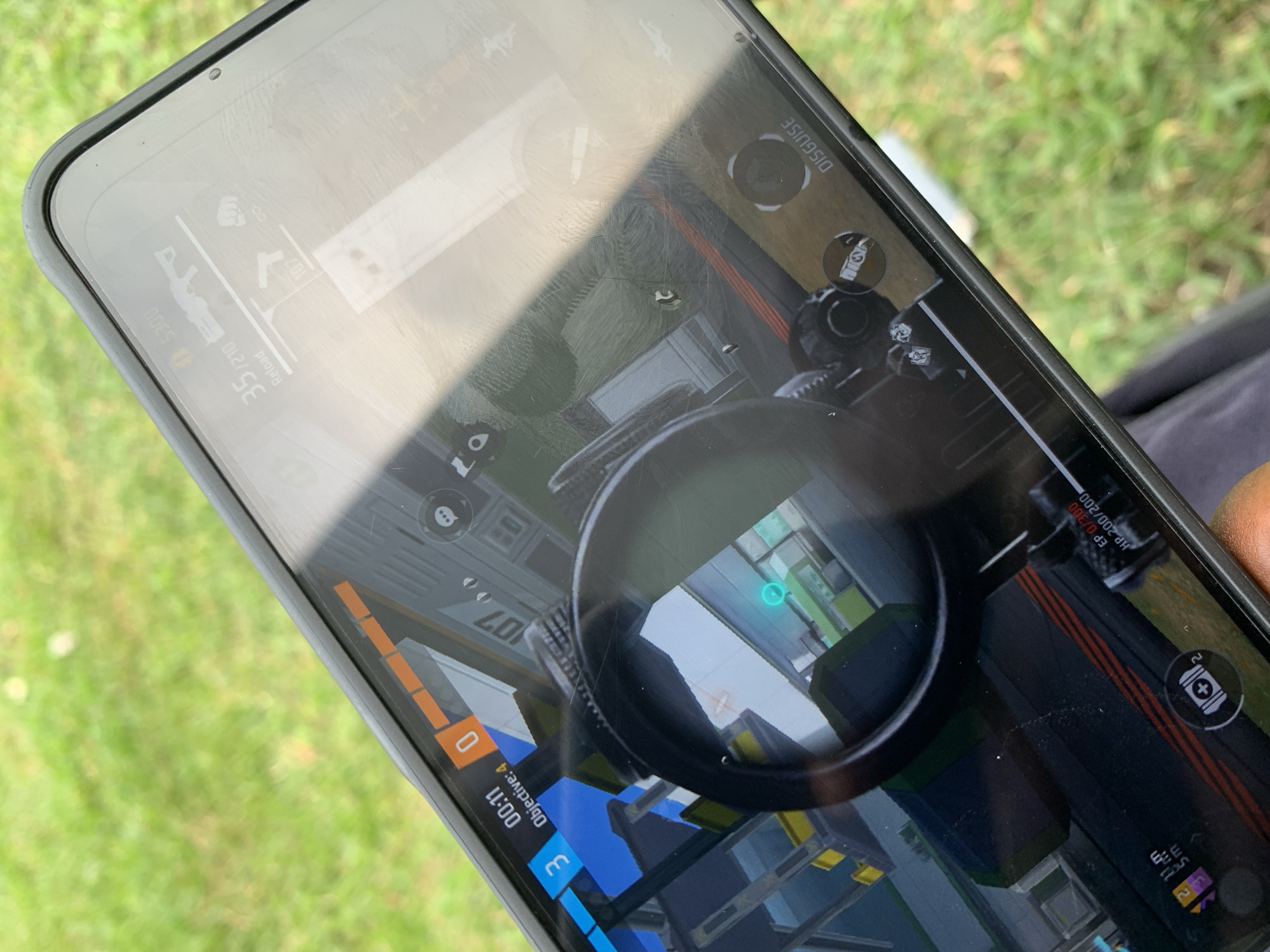 A close-up shot shows a smartphone displaying a first-person shooter game. The screen shows gameplay elements such as a heads-up display (HUD) with health and ammo counts, objective markers, a reload indicator, and a mini-map. A scope is visible, focusing on an in-game environment with geometric structures and lighting. The game appears to be set indoors or in a structured environment. The phone itself has a protective case. The background, blurred, suggests an outdoor setting with green grass. There is no evidence to identify the location as Sangotedo, Nigeria. The image does not show any people, interactions or actions, only subjects and objects.
