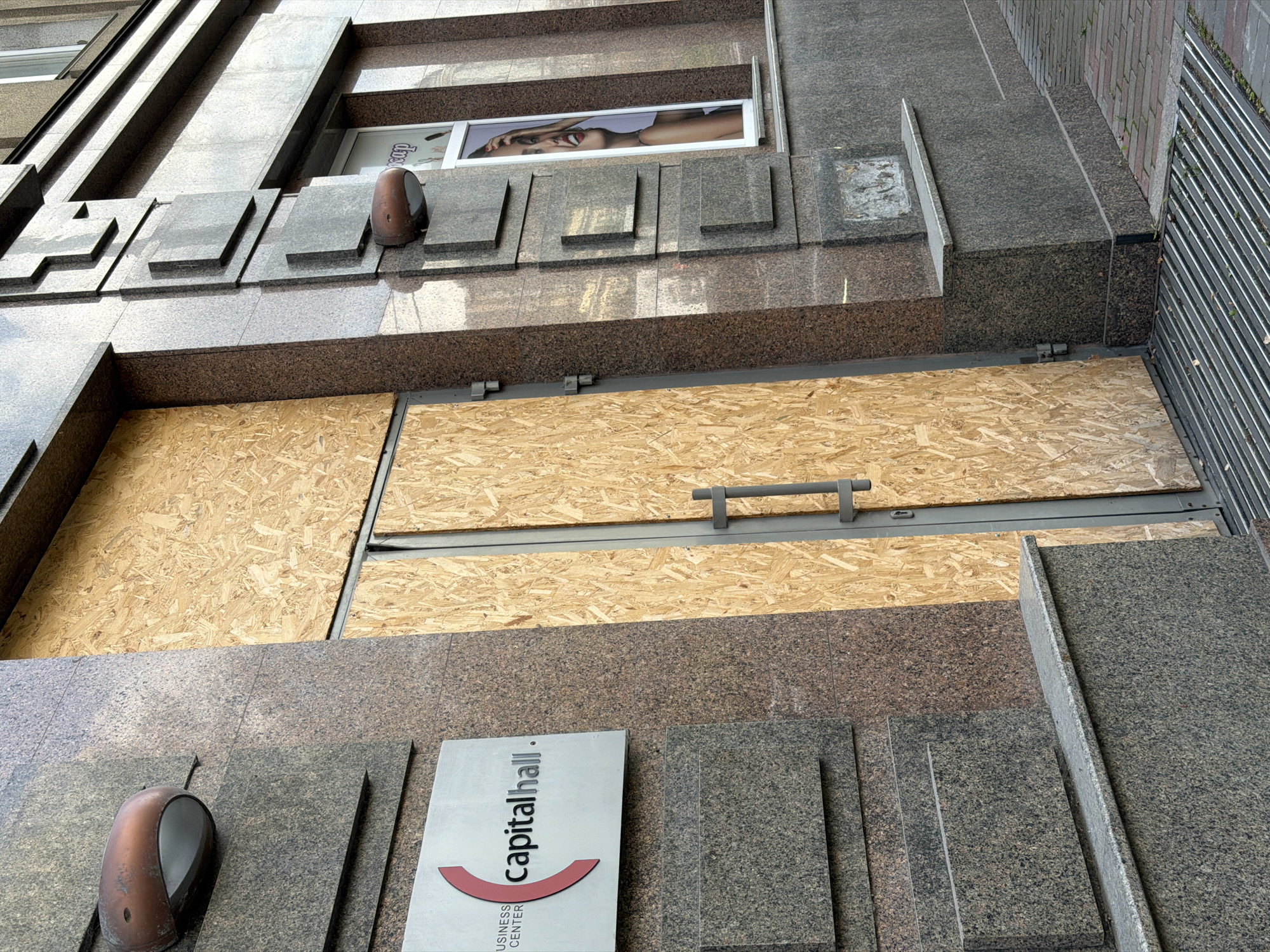 A medium shot captures the exterior of the Capital Hall Business Center in Kyiv, Ukraine. The main entrance is boarded up with plywood, featuring a gray metal door frame and a handle. To the left of the entrance, a white sign with red and gray lettering reads "BUSINESS CENTER capitalhall." The building's facade is clad in polished granite with a speckled brown and gray pattern, interspersed with darker gray rectangular blocks that create a stepped architectural detail. Two vintage-style copper wall lamps are affixed to the granite. To the right of the entrance, a window displays a poster of a smiling woman with her hand near her face. The ground in front of the building is paved with brick and features a metal grate drain.