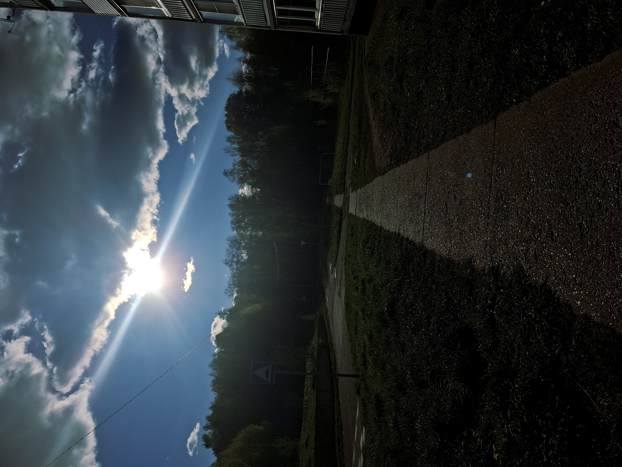 The image depicts an outdoor scene in Марковский, Russia. A pathway, likely made of gravel or small stones, runs vertically through the right side of the image. The pathway is shadowed on its right side, suggesting a light source on the left. Vegetation, including grass and trees, surrounds the pathway. In the background, a dense forest is visible. On the left side of the image, the sky is visible with bright sunlight and some clouds. A building is partially visible at the top of the image. A triangular traffic sign is visible near the bottom of the image.