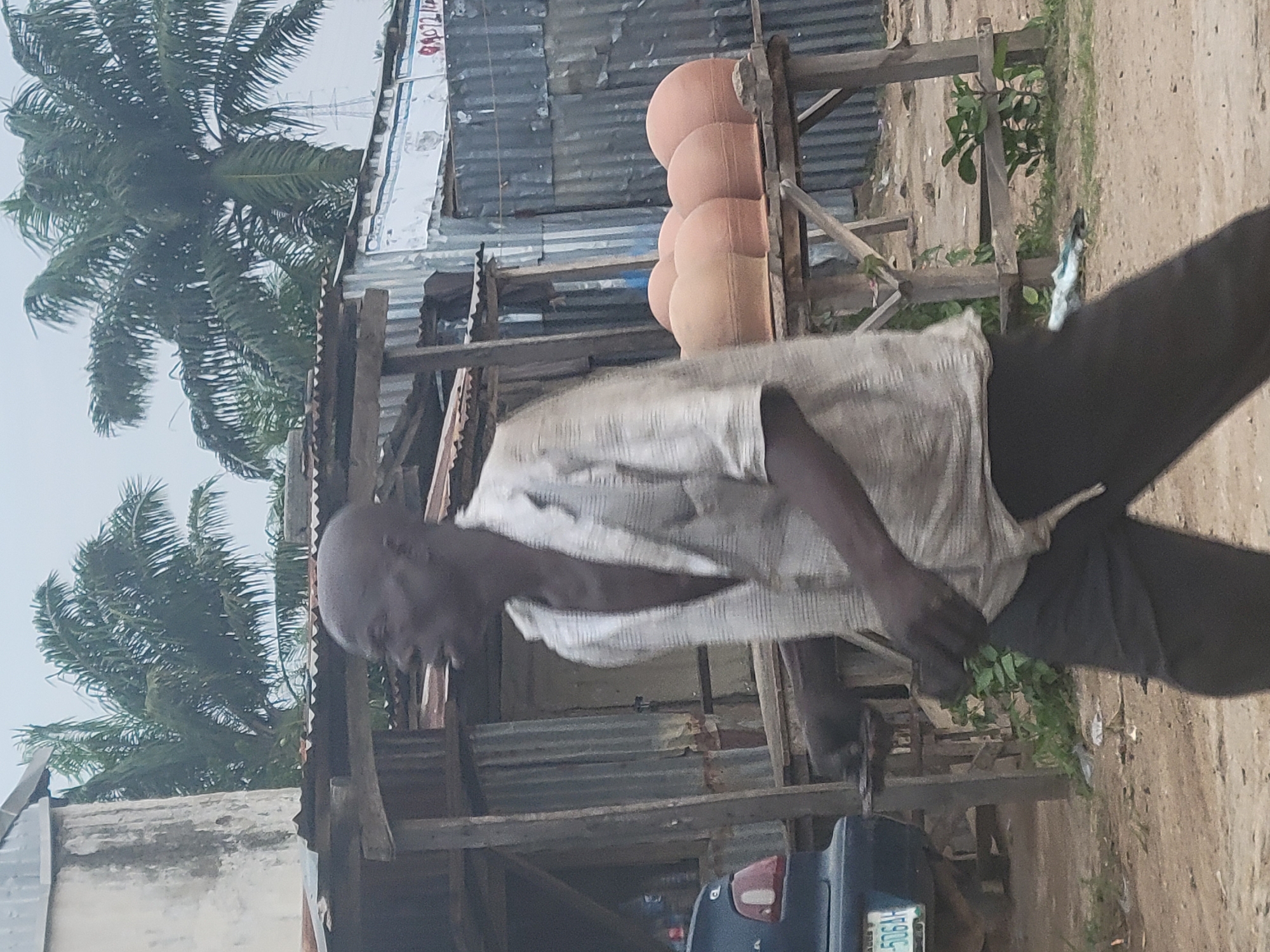 The image depicts an outdoor scene in Kwamba, Nigeria. A man, appearing to be of African descent and with a bald head, walks towards the left of the frame. He is wearing a light-colored, checkered shirt with the collar open, and dark pants. He is in motion, with his right arm slightly raised and his left hand down by his side. The man is walking past a wooden stand that displays several large, terracotta pots, arranged in a cluster. Behind the man and the stand, a corrugated metal wall is visible, suggesting a utilitarian or market setting. Palm trees can be seen in the background, indicating a tropical or sub-tropical environment. A dark-colored car is partially visible on the left side of the image, with a license plate that reads "506AH". The ground appears to be sandy or dirt. The lighting is overcast, suggesting a cloudy day. The overall atmosphere is one of everyday life and commerce in a Nigerian city.