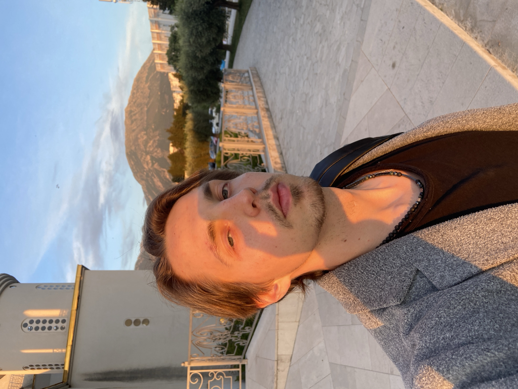 A medium-shot selfie of a young man with light brown hair, wearing a grey jacket over a dark brown shirt and a beaded necklace, with a goatee. He is standing outside, with a white building to his left and a mountain in the background. The photo is taken during golden hour, casting a warm light on his face. The location is the city of Bar, Montenegro, based on the presence of a distinctive Orthodox church tower with small rounded windows. A low stone wall with metal railings is visible in the foreground near the building. Paved ground extends from the wall. Behind the man, there are trees and other vegetation. The sky is blue with light clouds.