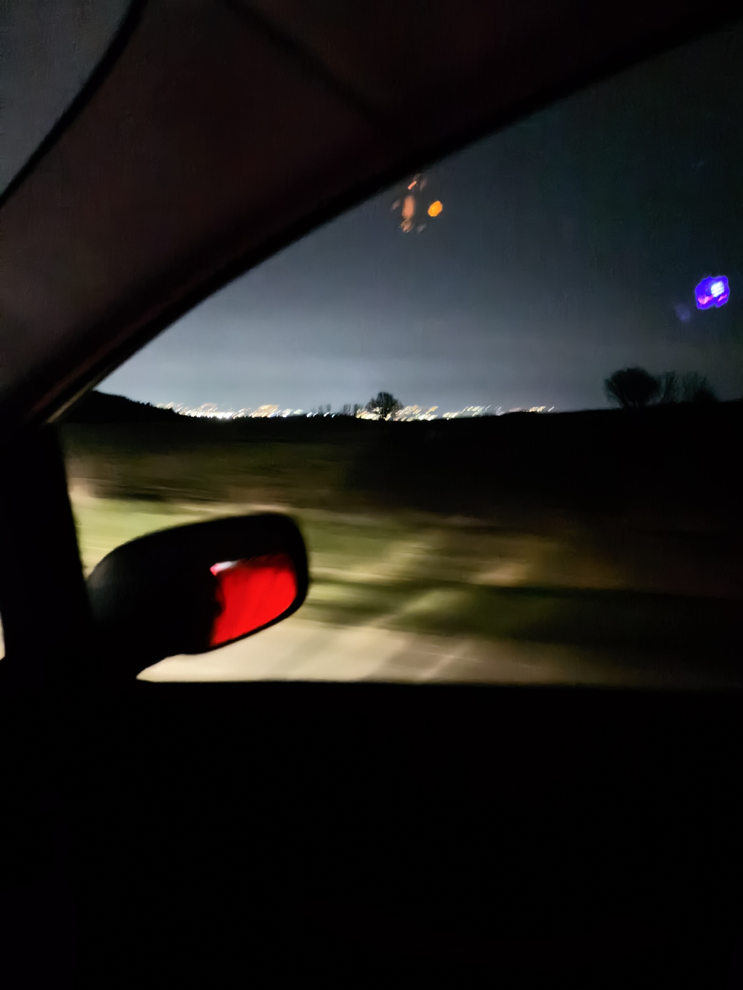 The view from inside a car at night. A blurred landscape with lights in the distance is visible through the window. The car's side mirror is visible with a red reflection. The location appears to be outside of a city, possibly on a road leading away from city lights. The exact location within Unknown, Unknown cannot be determined from this image.