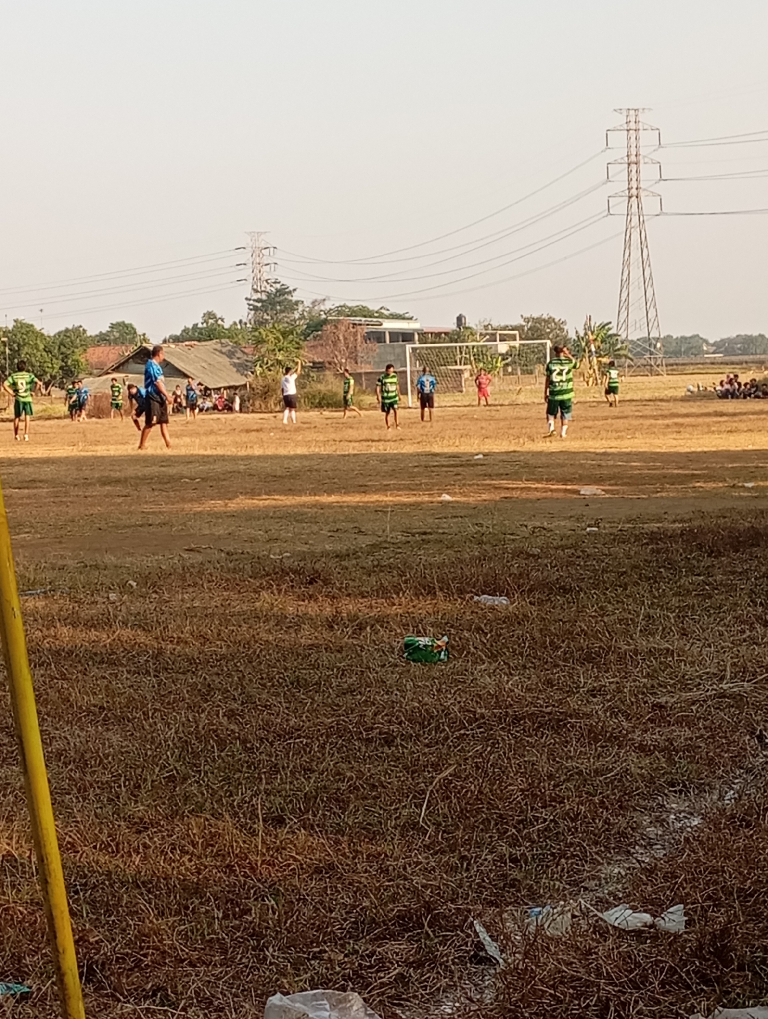 The image depicts a soccer game in progress on a dry, grassy field under a clear sky. Several individuals, some wearing green and white soccer jerseys and others in blue, are scattered across the field, suggesting an active match. A soccer goal is visible in the background. The environment includes a few low-rise buildings with corrugated roofs, trees, and utility poles with power lines stretching across the sky. The ground is covered with dry, brown grass and scattered debris, including plastic bags and bottles. In the foreground, a portion of a yellow pole is visible on the left side of the frame. The scene appears to be in an outdoor, possibly rural or semi-urban area, consistent with locations such as Kecamatan Haurgeulis, Indonesia.