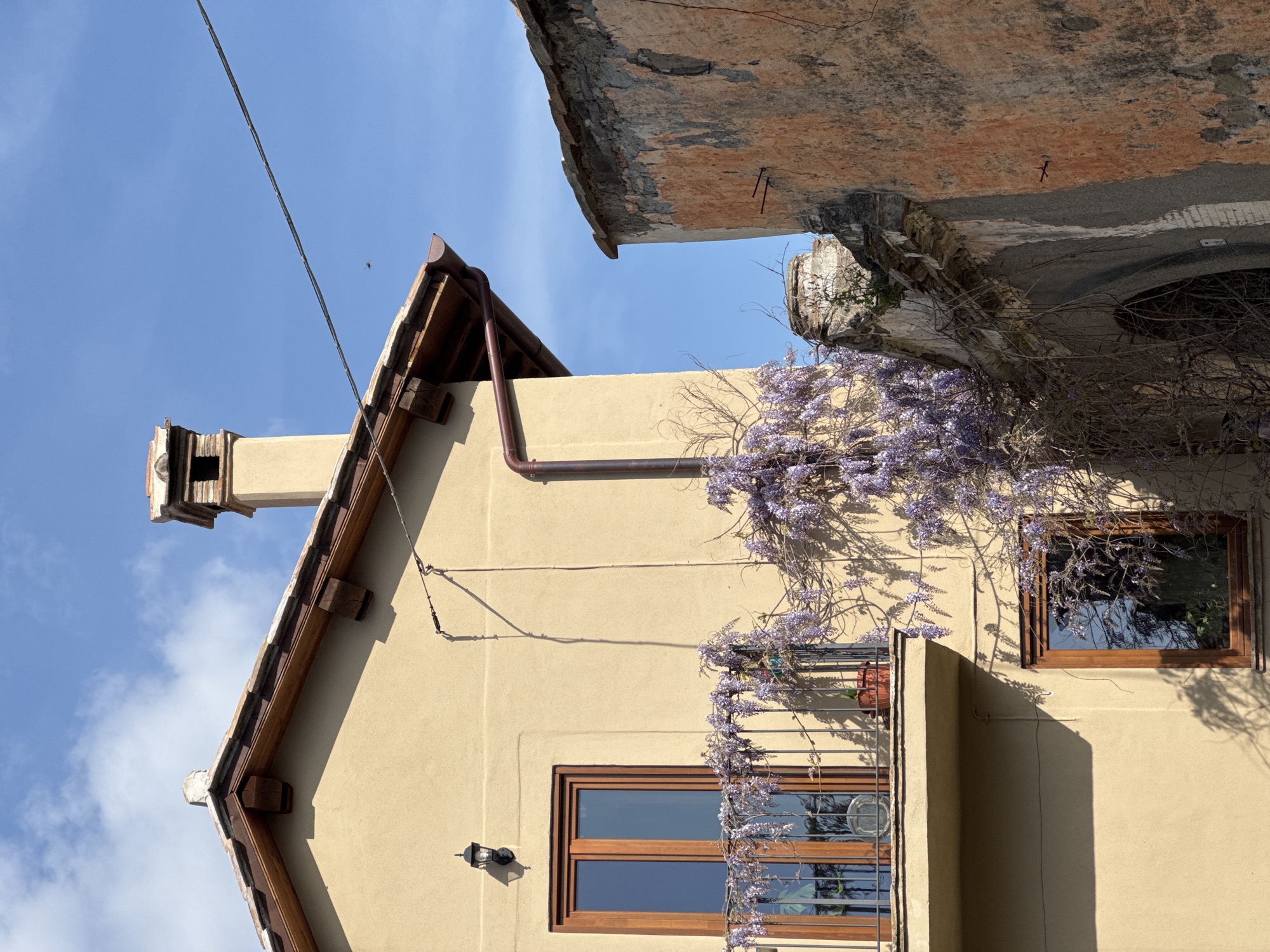The image depicts the exterior of a building in Castelnuovo di Porto, Italy, characterized by its light tan facade and a sloped roof with dark brown trim. A chimney protrudes from the roofline. The building features windows with wooden frames and a small balcony with a wrought-iron railing. Cascading wisteria vines adorn the building's exterior, adding a touch of color and natural texture. Above and to the right, a section of an older building is visible, with crumbling stucco and exposed brick, suggesting age and decay. The sky is blue with scattered clouds, and there is a cable wire crossing the scene.