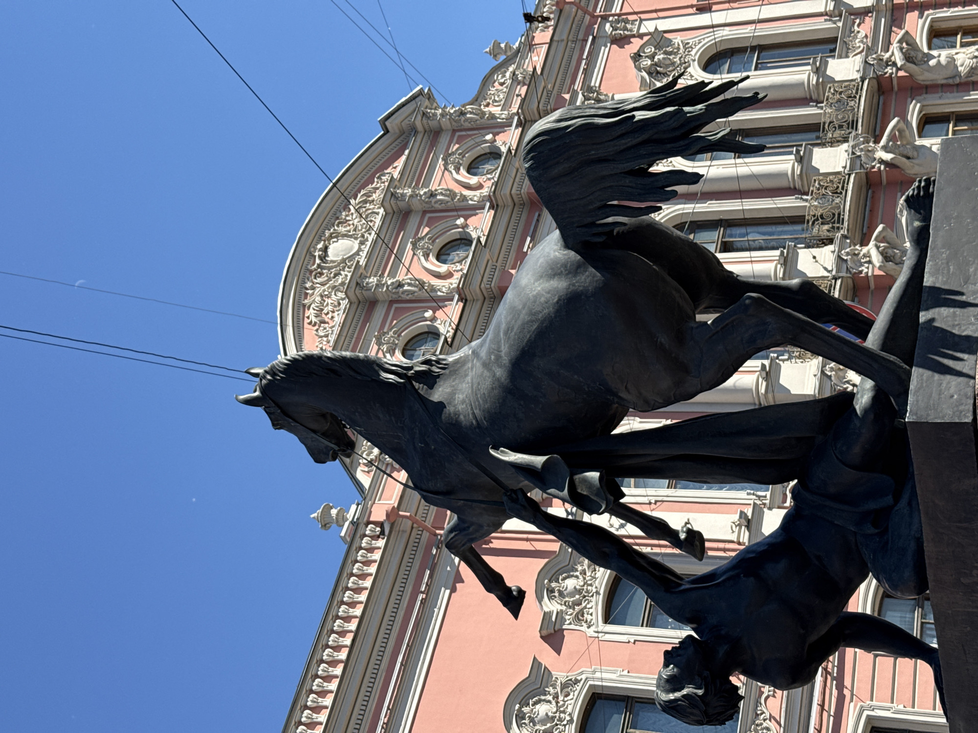 This media file features a dramatic, low-angle shot of a bronze sculpture in Saint Petersburg, Russia. The sculpture depicts a winged horse, possibly Pegasus, in mid-flight, with its mane and tail flowing as if caught in a strong wind. Beneath the horse, a partially visible human figure appears to be struggling or perhaps guiding the powerful creature. The dynamic poses of both the horse and the human suggest a moment of intense action or struggle.
The background is dominated by a grand, ornate building with a distinctive pink facade and intricate white stucco detailing around its windows and along the roofline. The architecture is elaborate, featuring curved window frames and decorative elements that suggest a historical or neoclassical style. The clear blue sky above, with a few faint wisps of clouds or dust motes, indicates fair weather and likely daytime, possibly late morning or early afternoon due to the bright sunlight casting shadows.
The presence of overhead wires suggests an urban setting, consistent with Saint Petersburg. The overall mood is one of grandeur and perhaps mythological epic, conveyed by the imposing sculpture and the majestic building. The perspective emphasizes the scale and power of the artwork. No visible text is present in the image.
