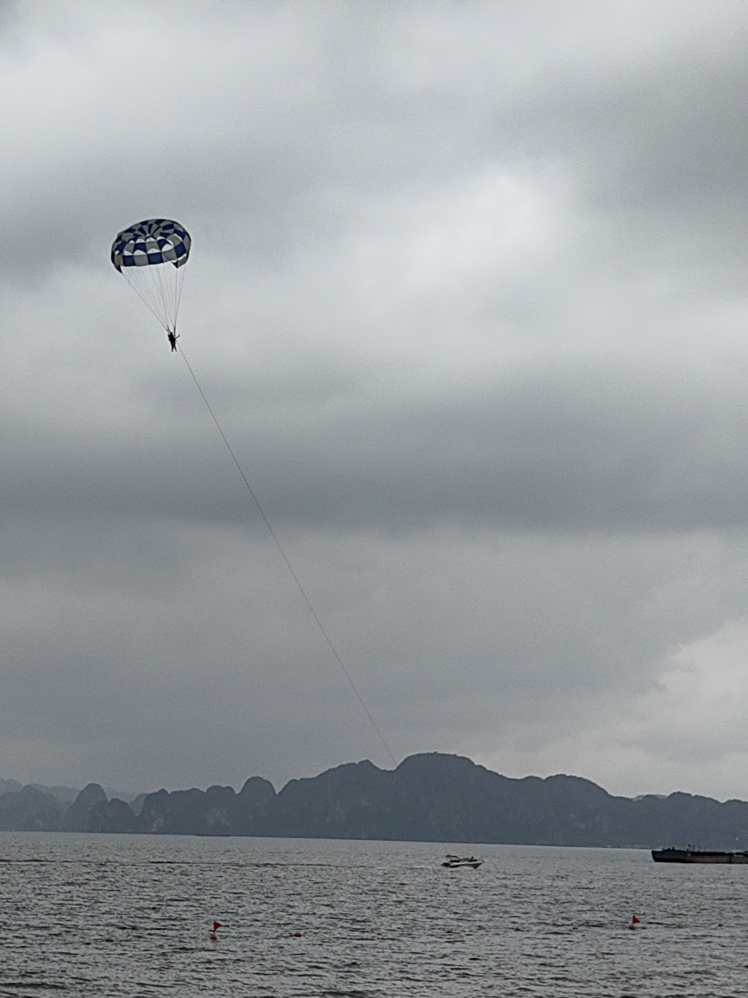 The image depicts a parasailer suspended in the air over the waters of Bai Chay Ward, Vietnam, under an overcast sky. The parasail is patterned with blue and white checks. Below the parasailer, the landscape includes a body of water with several distant rocky islands and a large, dark mountain range. There is a small motorboat on the water's surface and a larger vessel farther away. Several red markers are also visible in the water closer to the foreground. The sky is covered in thick grey clouds.