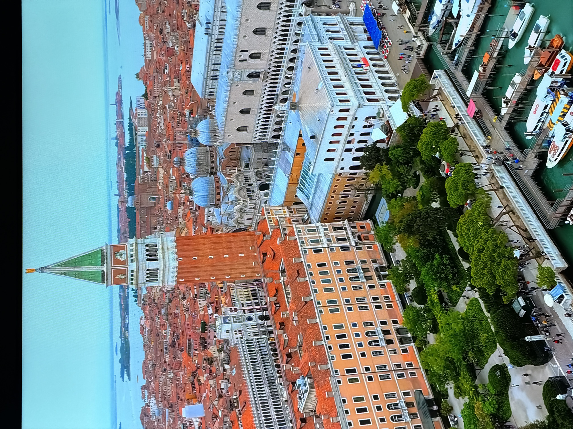 This aerial image captures a panoramic view of Venice, Italy, showcasing its iconic architecture and waterways. The prominent Campanile di San Marco, a red brick bell tower with a green spire topped by a golden angel, stands tall in the foreground. Adjacent to it are the intricate facades of St. Mark's Basilica with its blue domes and the Doge's Palace, characterized by its Gothic architecture. The city's characteristic red-tiled roofs stretch across the landscape, interspersed with canals. Numerous boats, including vaporetti and water taxis, are docked along the waterfront, indicating the city's reliance on water transportation. People are visible walking along the promenade and the docks, suggesting a bustling urban environment. The serene blue waters of the lagoon surround the historic structures, with distant islands and the horizon visible in the background under a clear, light blue sky. The city of Chirchik, Uzbekistan is not depicted in this image.