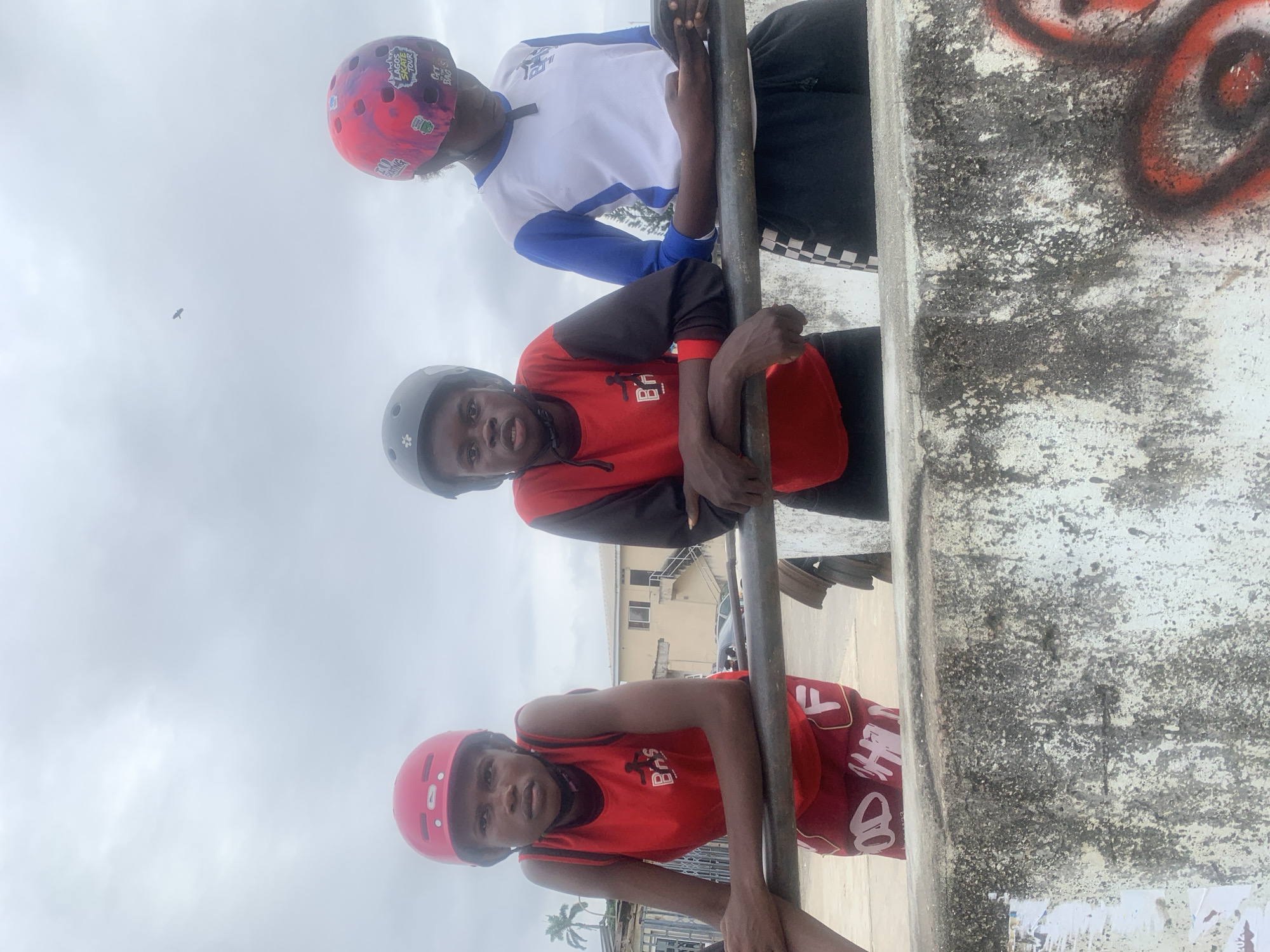 Three young people, two wearing red helmets and one a colorful helmet, are standing behind a rusty metal railing outdoors. The person on the left, with a red helmet and red sleeveless shirt, rests their arms on the railing. The person in the middle, wearing a black helmet and a red long-sleeved shirt with black sleeves, leans their arms on the railing. The person on the right, with a multicolored helmet, wears a white and blue long-sleeved shirt and is looking down at a mobile phone held in their hands. They are all wearing dark-colored pants. In the background, there is a building, some parked cars, and palm trees, suggesting an urban setting. The sky is overcast. The graffiti on the concrete structure in the foreground indicates a public or recreational space. This scene is in Alagbede, Nigeria.