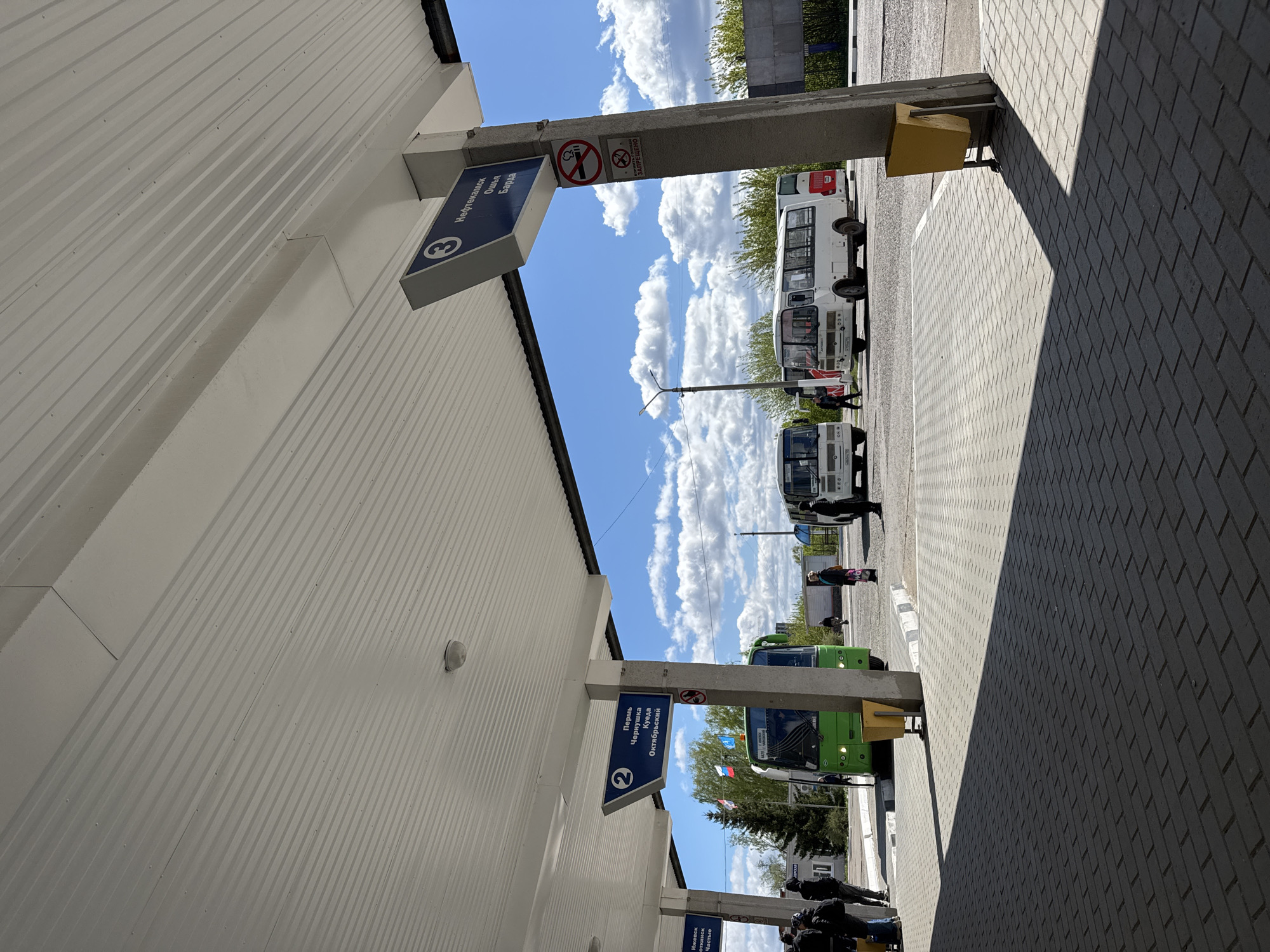 The image shows a bus station in Chaikovsky, Russia. Several buses are parked at numbered bays, and passengers are waiting or boarding. The sky is blue with fluffy white clouds. No smoking signs are visible.