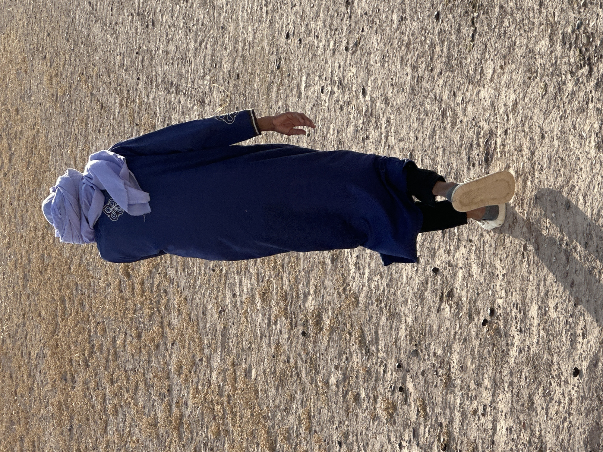 A man wearing a dark blue traditional Moroccan djellaba and light blue cheich is lying on a flat, dry, barren landscape. The ground appears to be a mixture of sand and dry vegetation. The man's head is wrapped in the cheich, and his face is not visible. He is also wearing black pants, gray socks, and white shoes. The location is presumed to be near the city of Sidi Badhaj, Morocco.