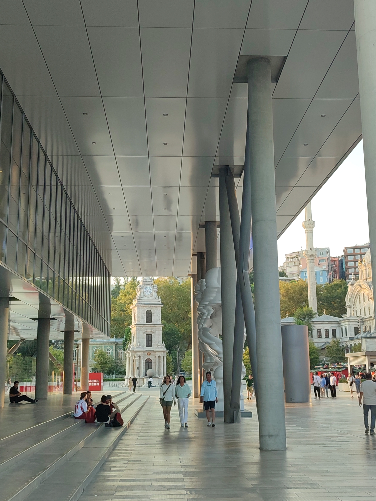 The image depicts an outdoor scene in the Beyoğlu district of Istanbul, Turkey, likely in the vicinity of the Tophane neighborhood given the architecture. In the foreground, a modern building with a large, reflective glass facade runs along the left side of the frame, extending overhead and supported by large, cylindrical concrete pillars. The ceiling of this modern structure is made of metallic panels. A wide, stepped seating area leads from the foreground towards the background.
On the steps, several individuals are seated, appearing to be engaged in conversation. One person sits alone on the lower left, facing away from the camera. Further up the steps, a group of four individuals are gathered. To the right of the stepped area, a paved walkway is visible, where several people are walking. Two women are walking towards the camera, dressed in casual attire. Further in the background, a group of people are seen standing near what appears to be a marketplace or event area with stalls.
In the middle ground, a distinctive clock tower, likely the Tophane Clock Tower, stands prominently. Behind the clock tower, trees are visible, with more urban architecture in the distance. A minaret of a mosque also rises into the sky to the right of the clock tower, indicating the presence of religious buildings. The overall atmosphere suggests a public urban space with a blend of modern and historical architecture, bustling with people. The lighting indicates it is daytime, with the sun casting shadows.