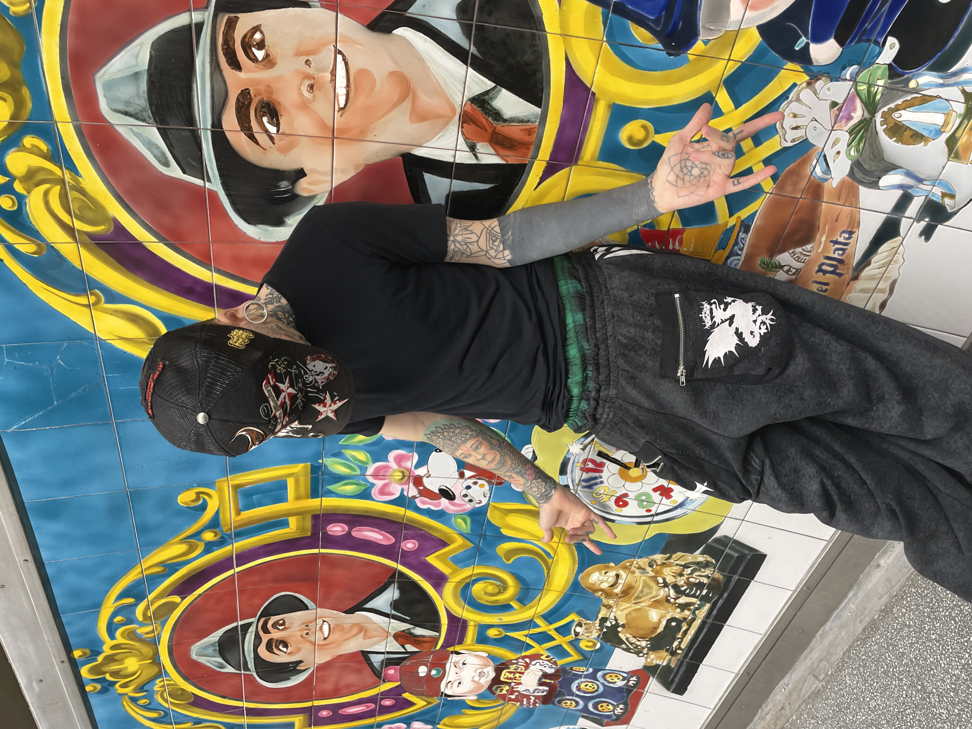 A person with extensive tattoos stands on a tiled mural. The mural depicts various images including portraits of men in suits, clocks, and figures. The tiles are arranged in a grid pattern with blue, gold, and red colors. The person wears a black t-shirt, dark grey sweatpants with a dragon graphic, and a black baseball cap. They make a hand gesture with both hands. The setting is in the city of Buenos Aires, Argentina.