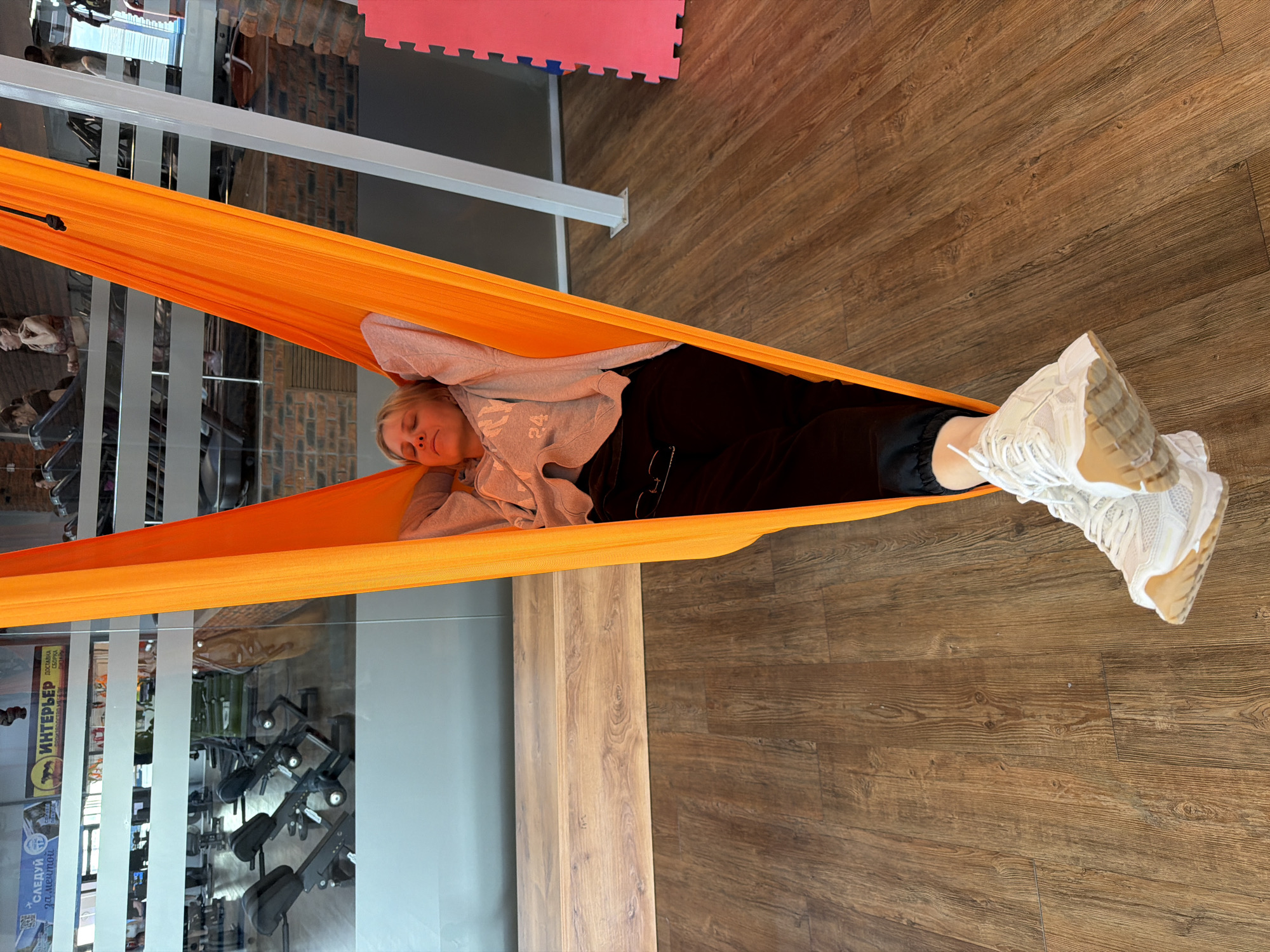 The image, originally oriented vertically, captures a serene scene in what appears to be an indoor fitness studio or gym in Chaikovsky, Russia. A young woman with blonde hair is seen relaxing horizontally in a bright orange aerial hammock. She lies with her head resting on her hands, eyes closed, displaying a peaceful and calm expression. She is dressed in a light grey long-sleeved top and dark pants, with her feet, clad in chunky white sneakers with tan soles, extending out from the hammock.
The floor beneath the hammock is made of dark brown wooden planks. The background reveals reflections in glass panels, showcasing various pieces of gym equipment, including what appear to be rowing machines and other exercise apparatus, suggesting a workout area. White structural beams and sections of red and blue interlocking foam mats are also visible on the floor in the background, reinforcing the gym setting.
Visible text in the background reflections includes Russian words such as "ИНТЕРЬЕР" (INTERIOR), "ДОСТАВКА" (DELIVERY), and "СБОРКА" (ASSEMBLY). Also present are "СЛЕДУЙ 2Д" (FOLLOW 2D) and a logo depicting a horse-like animal with the word "УРАЛ" (URAL). The lighting is bright and even, consistent with an indoor daytime setting, contributing to the tranquil atmosphere.