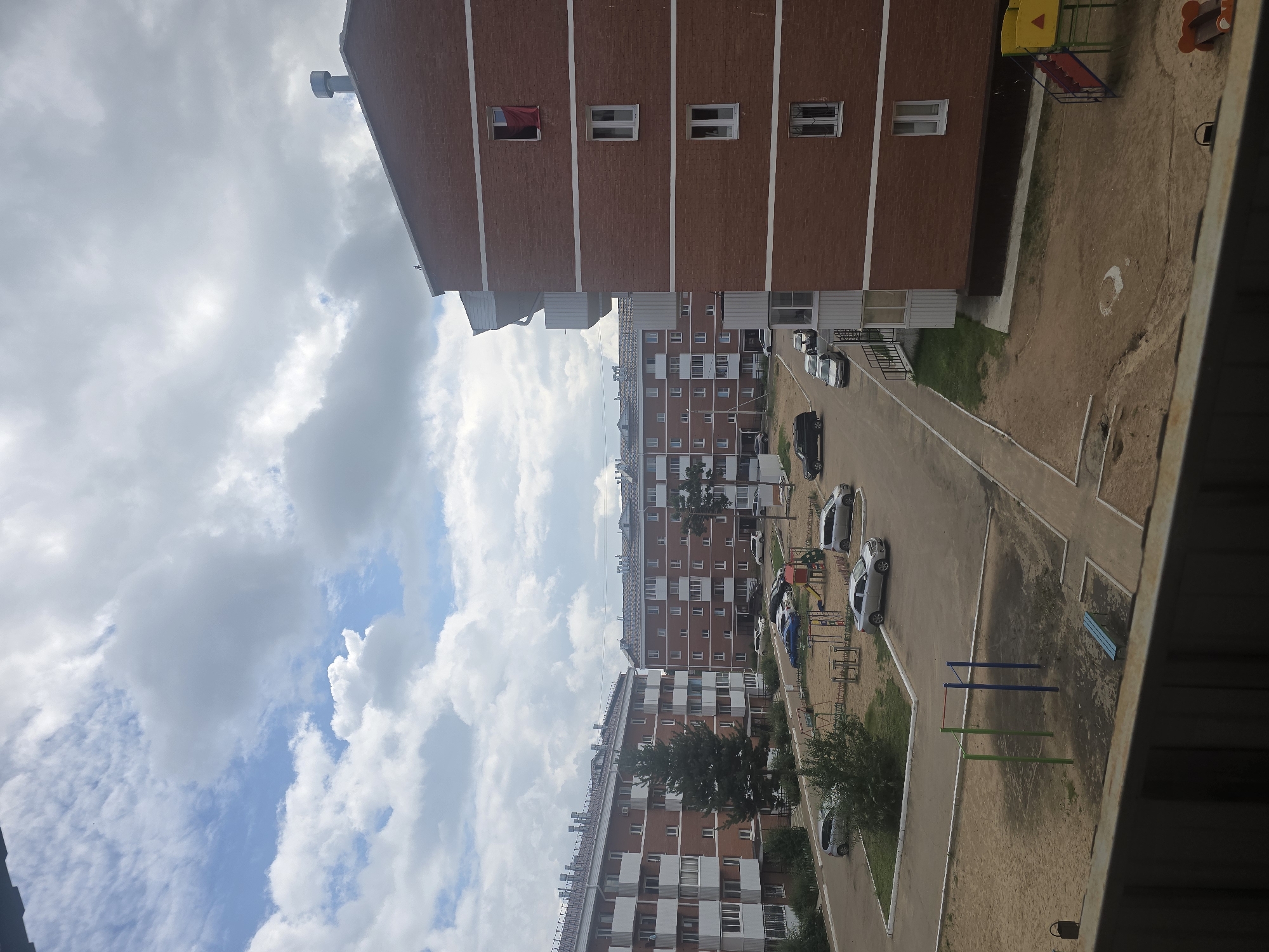 The image captures a view of a residential courtyard in Ulan-Ude, Russia, under a partly cloudy sky. The scene is dominated by several multi-story brick apartment buildings, typical of Soviet-era or post-Soviet urban architecture. The buildings are reddish-brown with white trim around the windows and balconies.
In the foreground, a balcony railing is visible, suggesting the photo was taken from a window or another balcony. The courtyard features a sandy playground with a slide and swings, along with exercise bars. A paved road or pathway winds through the courtyard, and several cars are parked. One building, on the right side of the frame, has a red cloth hanging from an upper window. Green trees and bushes are scattered throughout the courtyard, adding some natural elements to the urban landscape. The overall impression is one of a typical, functional residential area.