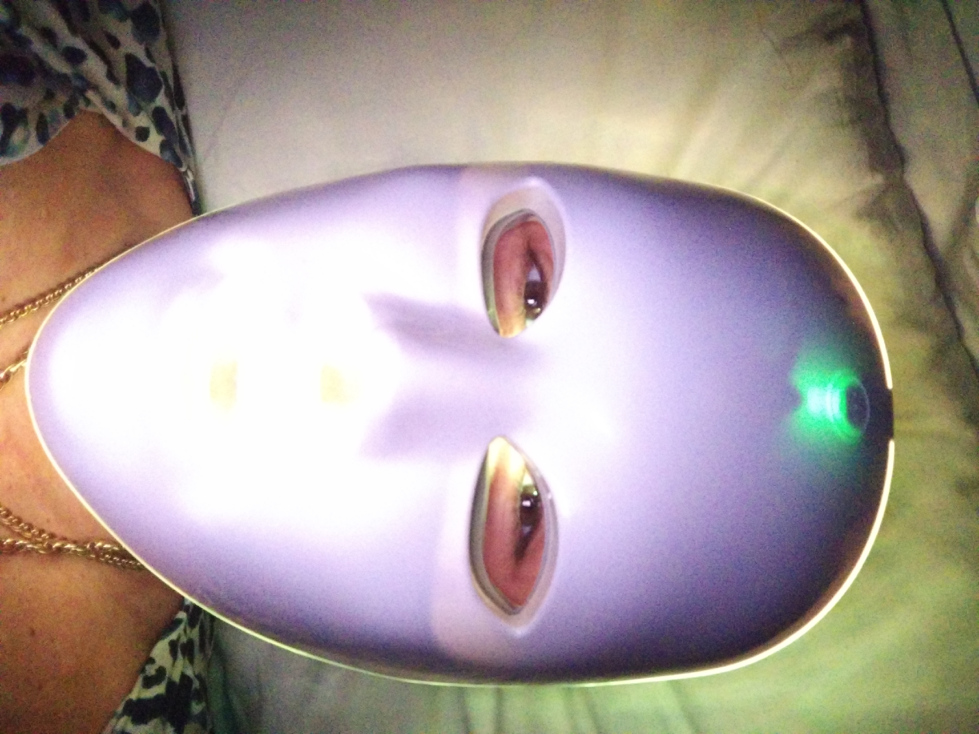 The image shows a close-up, eye-level, portrait-style photograph of a person wearing a solid, opaque, light purple mask. The mask covers the entire face and has two almond-shaped eye openings. A person's eyes, with brown irises and dark pupils, are visible through the openings. The mask has a smooth, curved surface with a subtle sheen. A bright green light emanates from a source above and behind the mask, casting a soft glow and creating highlights on the mask's surface. The background is blurred and appears to be a textured fabric, possibly a duvet or blanket, in muted tones of grey and green. A portion of the person's neck and upper chest are visible below the mask, where they are wearing a patterned top with a black and white, possibly leopard-print, design and a thick, gold-colored chain necklace. The image is overexposed in some areas due to the bright light source. The photograph was taken in Stara Zagora, Bulgaria.