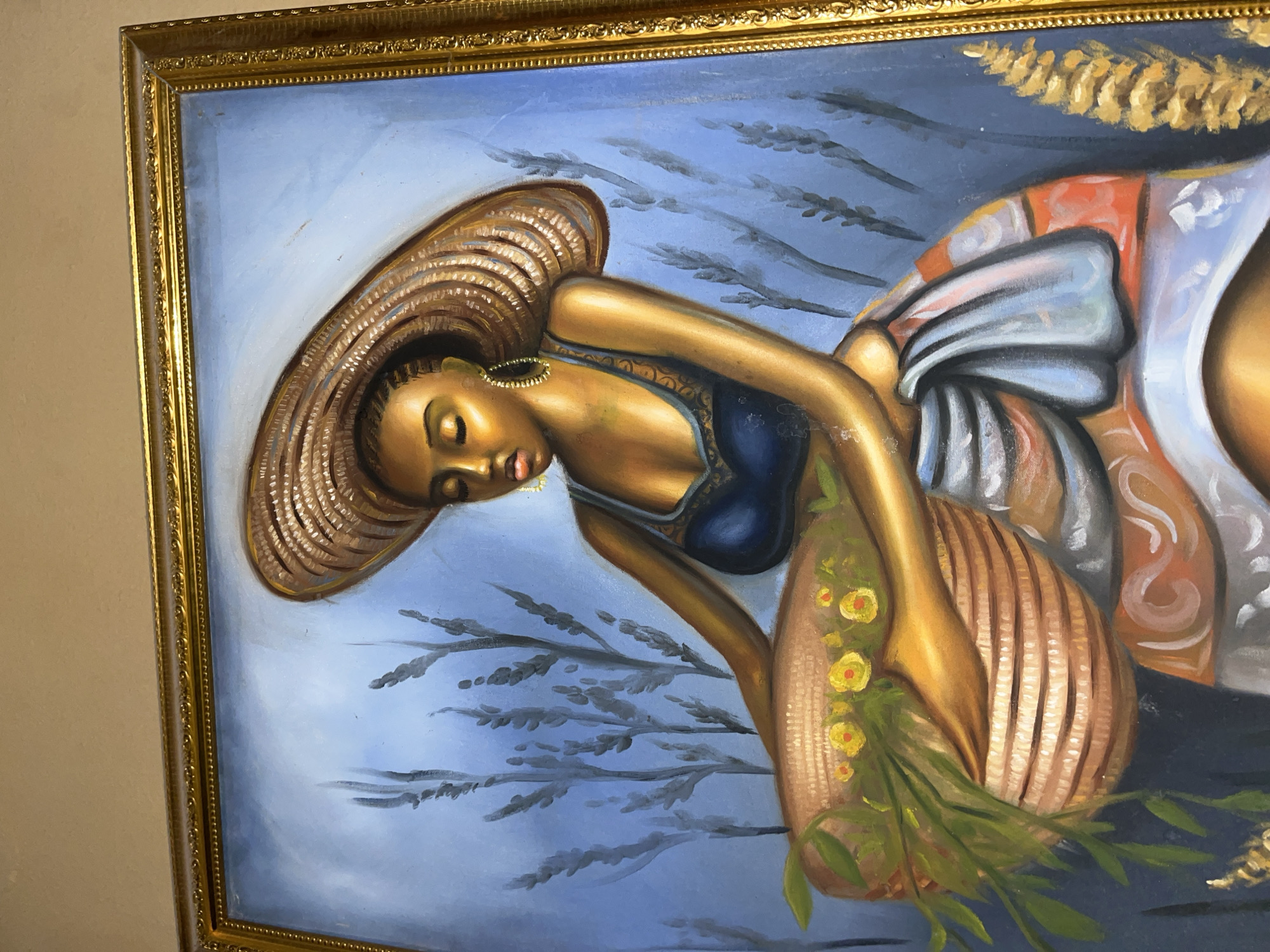 The image is a close-up of a framed oil painting. The painting depicts a Black woman wearing a wide-brimmed hat and a blue top. She is leaning over what appears to be a woven basket with flowers and foliage in it. The background is a hazy blue, with stylized depictions of what could be wheat or similar crops. The painting has a gilded ornate frame. There is no verifiable information available to place this painting or scene specifically in Madalla, Nigeria, or confirm its authenticity.