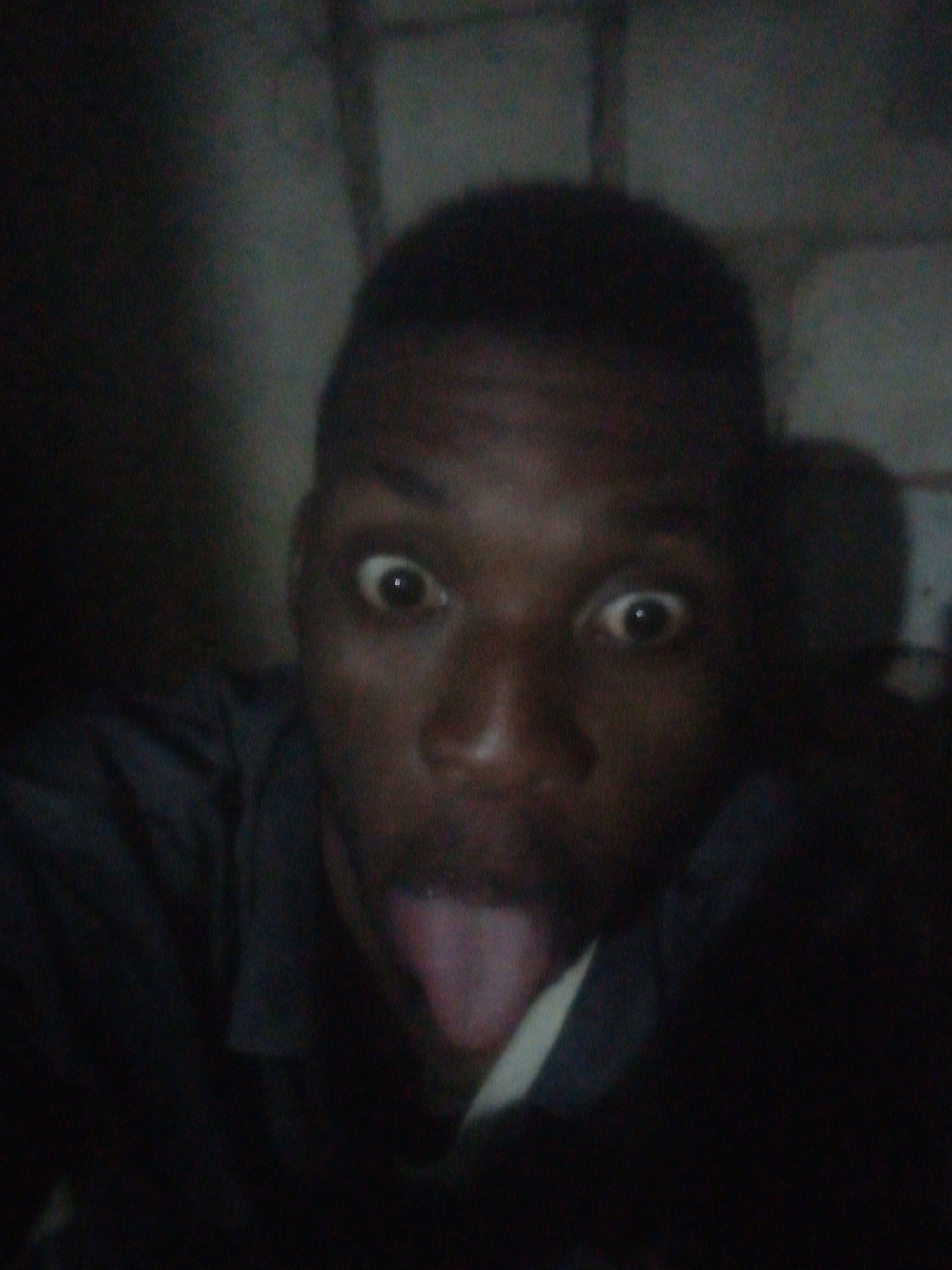 A close-up portrait captures a young man with dark skin and short, dark hair, positioned centrally in the frame. He is wearing a dark, collared shirt. The man is making a humorous or exaggerated facial expression, characterized by wide-open eyes with visible whites and dilated pupils, and his tongue sticking out from his mouth. The lighting in the image is very dim, casting the subject in significant shadow. The background is almost entirely dark and indistinct, suggesting an indoor setting with a plain wall or surface behind him, poorly illuminated. The overall impression is an informal, playful moment, possibly a selfie taken in low-light conditions. There is no visible text.