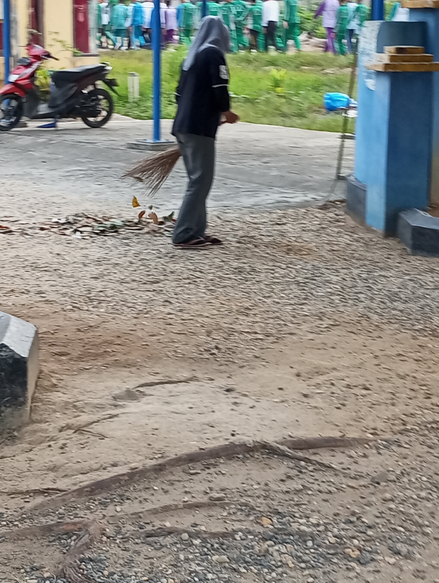 In Pematang Reba, Indonesia, a person wearing a hijab and a dark jacket with grey pants sweeps the ground with a broom made of twigs. The person is standing on a paved area with patches of sand and small stones. Behind the person, a red and black motorcycle is parked. In the background, several children in green and white uniforms are visible. To the right, a blue pillar and a blue trash can are present. The environment appears to be outdoors, possibly near a school or public facility.