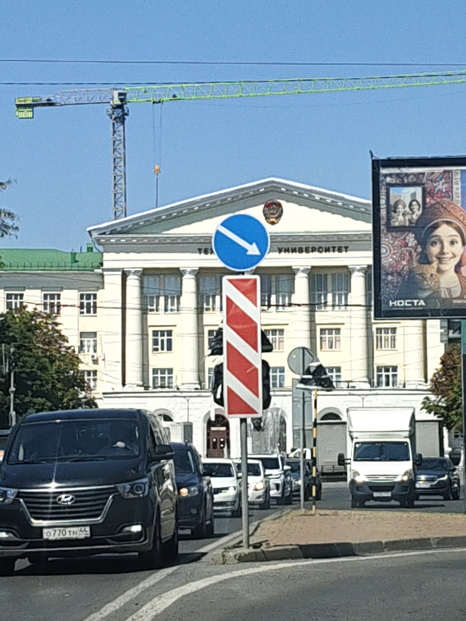The image captures a street scene in Rostov-on-Don, Russia, dominated by the imposing facade of the Rostov State Economic University, identifiable by the Cyrillic inscription "УНИВЕРСИТЕТ" (University) above its classical columns. A large construction crane with a green boom is visible in the background, suggesting ongoing development or maintenance. In the foreground, several vehicles are on the road, including a black minivan with a license plate reading "О770 TH 64", a white truck, and other cars. Traffic signs, including a directional arrow sign and a red and white striped sign, are present. A large advertisement poster featuring a woman in traditional attire and a cat is affixed to a building on the right side of the frame. The sky is clear and blue. The environment appears to be a bustling urban area.