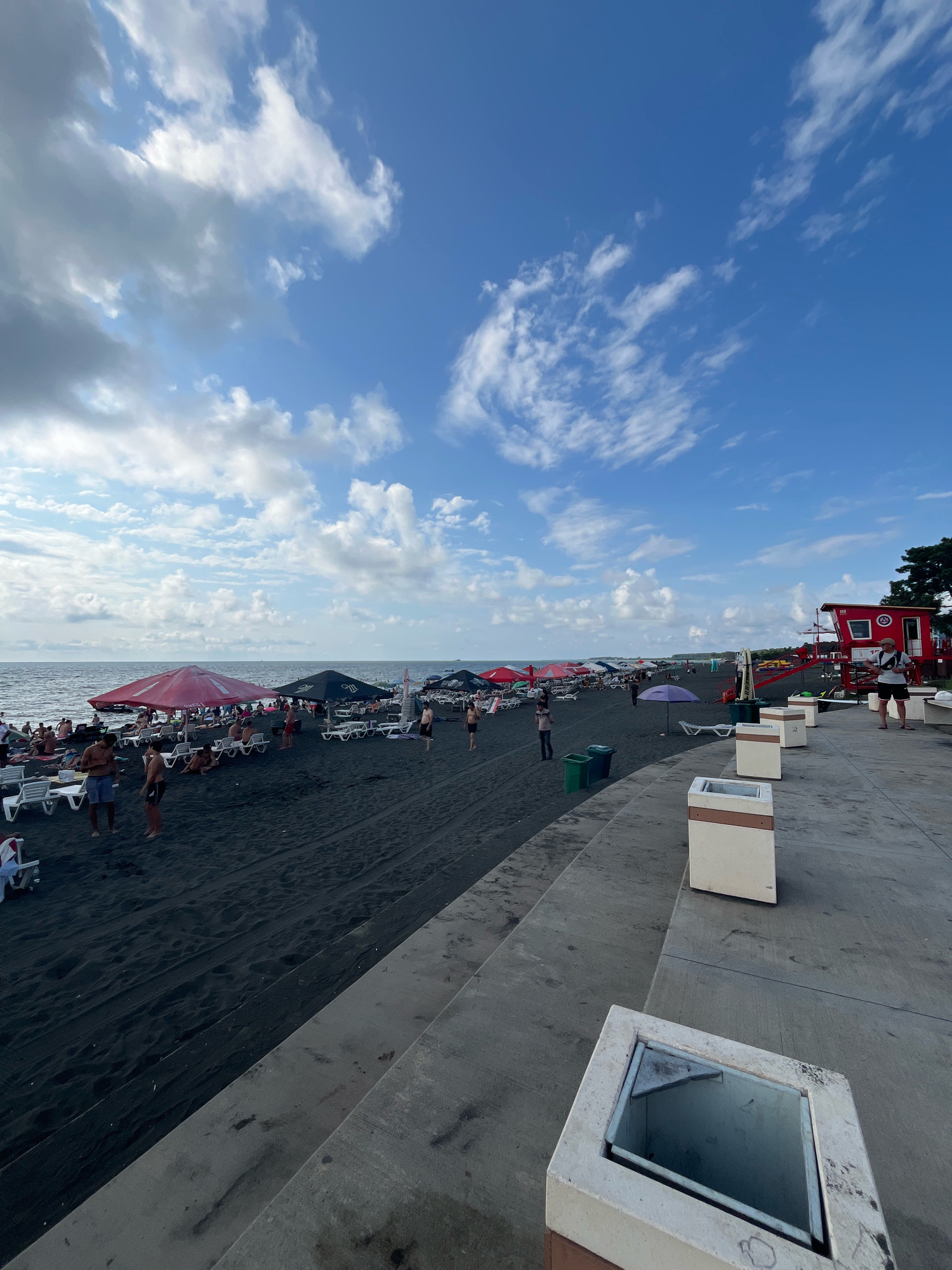 The image depicts a crowded black sand beach in Ureki, Georgia, under a partly cloudy sky. The beach is lined with numerous lounge chairs and umbrellas, indicating a popular tourist destination. Several people are visible, some sunbathing on the chairs, others walking along the shore or standing near the water. The sea stretches out to the horizon, with gentle waves lapping the dark sand. In the foreground, a concrete walkway with some planters and trash receptacles is visible, suggesting a developed promenade area. To the right, a red lifeguard station with the number "112" is prominent. The overall scene portrays a typical day at a bustling beach resort.