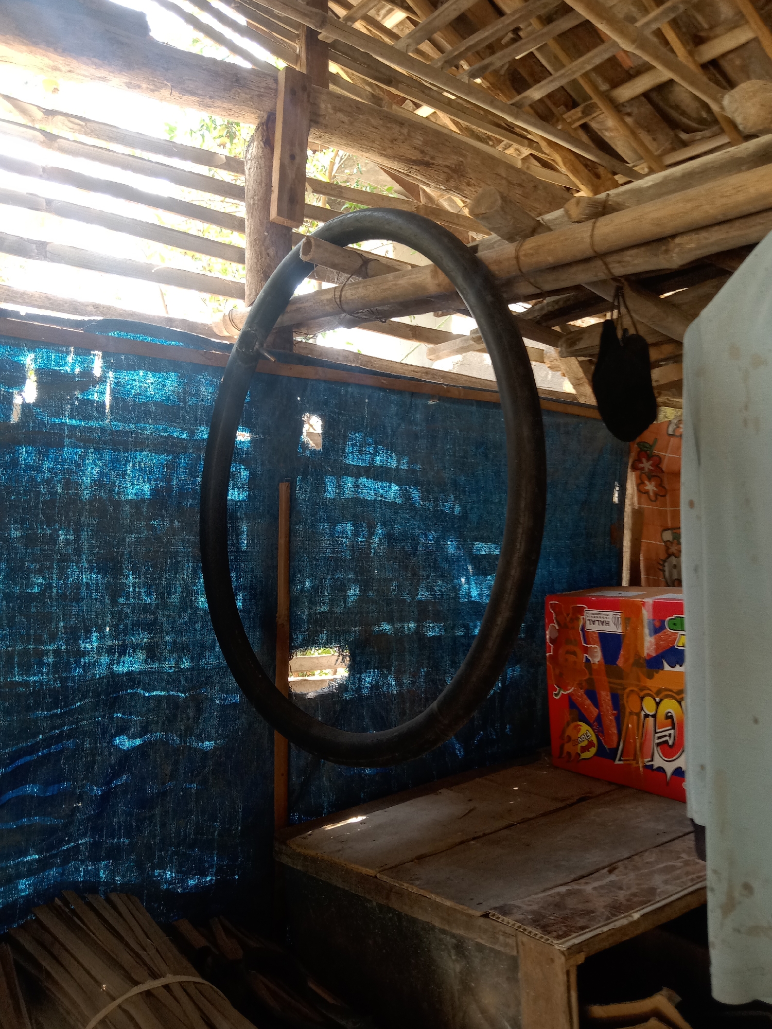 The image depicts the interior of a rustic structure, likely a shed or a simple workshop, in Stiaji, Indonesia. The ceiling is constructed from bamboo poles and translucent sheeting, allowing sunlight to filter through. A large, dark-colored hose or tube is suspended horizontally from the ceiling by rope. A blue tarpaulin serves as a wall covering on the left side of the frame. To the right, a wooden platform holds a brightly colored box, possibly merchandise, featuring text that includes "HALAL" and "INDONESIA." A white object, perhaps a piece of fabric or furniture, is partially visible on the far right. Bundles of dried palm leaves are stacked on the left side of the floor. The overall scene suggests a functional space used for storage or light industrial activity in a rural or semi-rural Indonesian setting.