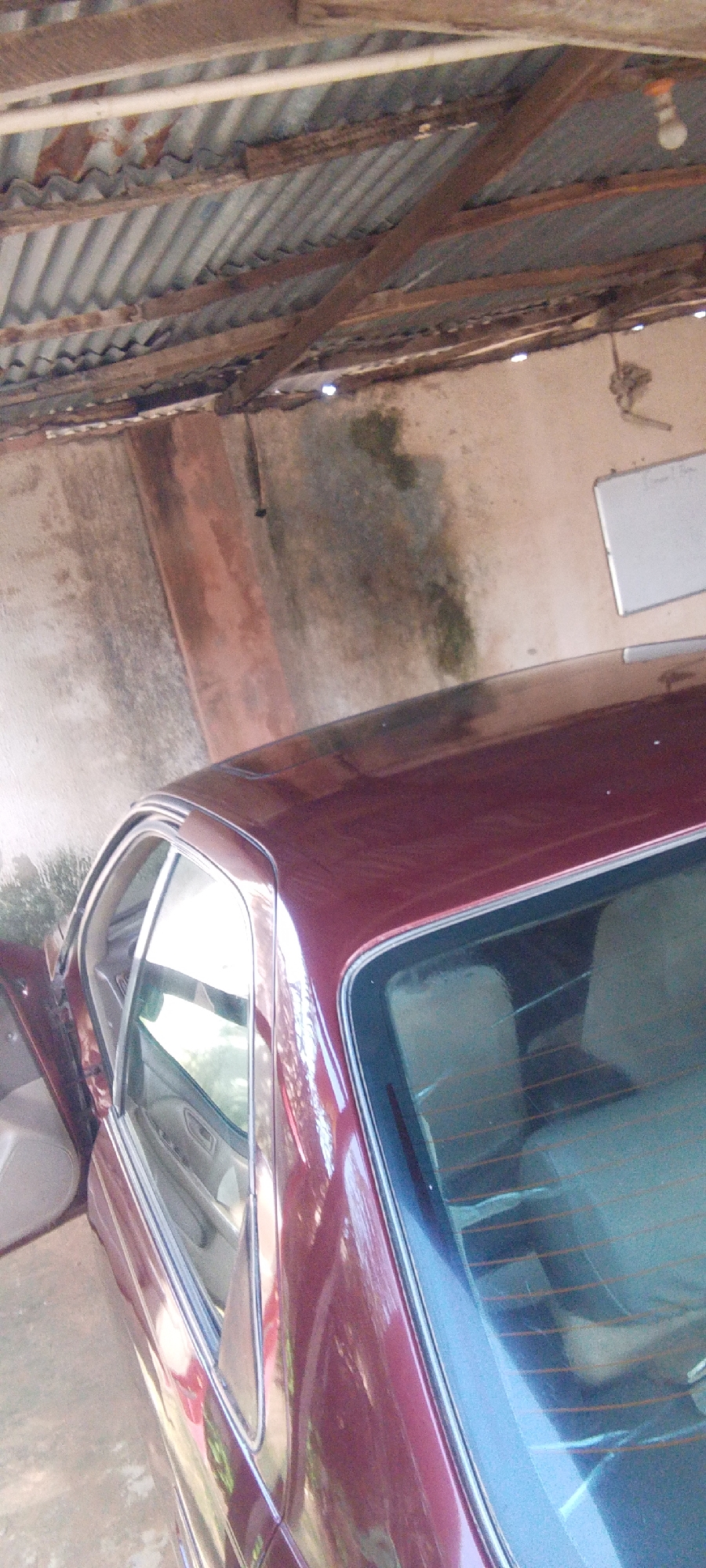 The image shows a maroon sedan parked in a covered area, possibly a garage or shed, with a corrugated metal roof supported by wooden beams. The walls appear to be concrete and are stained with what looks like water marks and green algae in places. A whiteboard is visible on the far wall. The open driver's side door reveals the car's interior. The scene is likely outdoors, given the lighting which suggests daytime. There are no people visible in the image. The focus is on the car and its immediate surroundings, hinting at a potentially unfinished or rustic structure. The location is identified as Madalla, Nigeria.