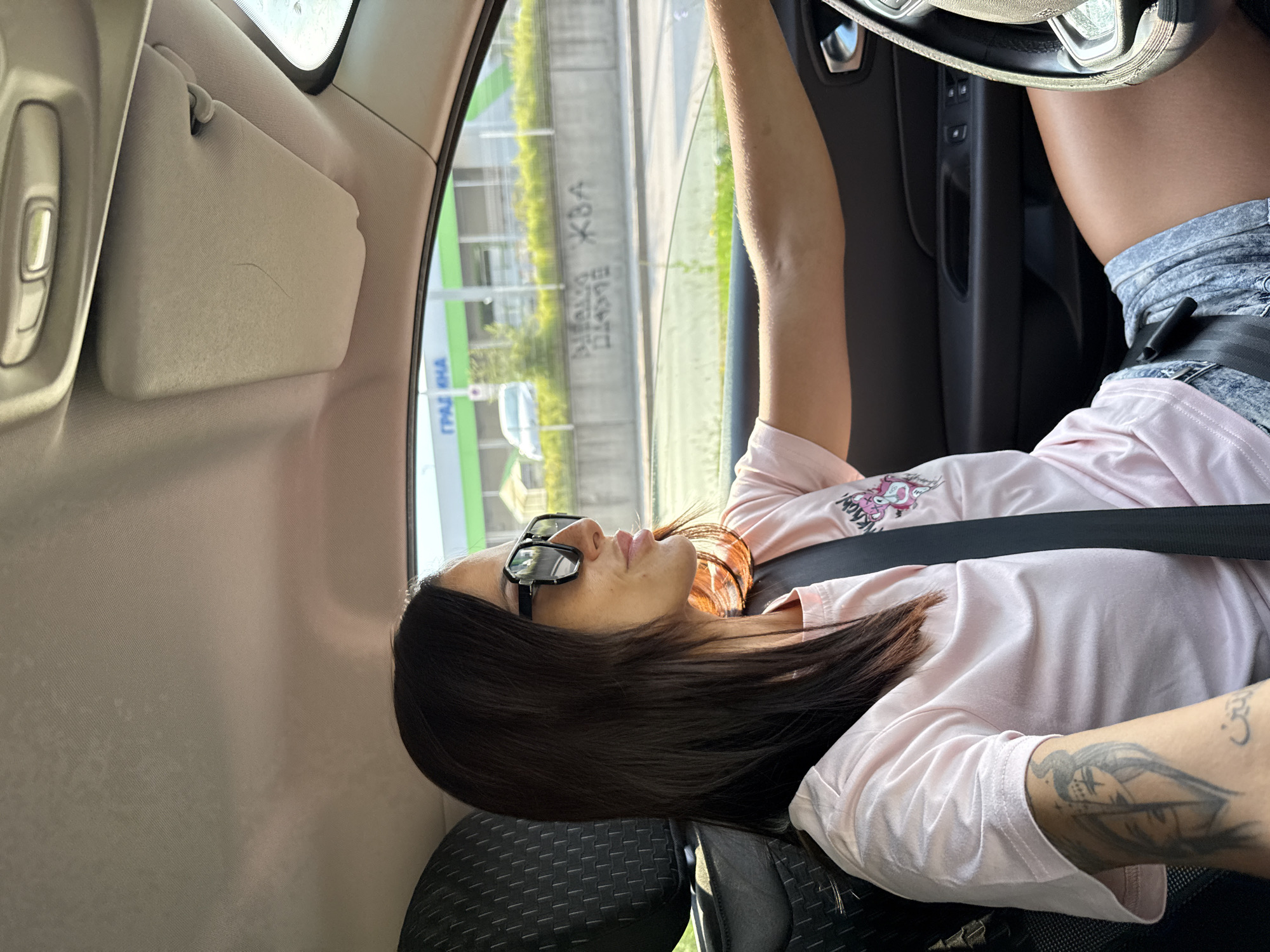 A woman with long dark hair and sunglasses is driving a car. She is wearing a pink t-shirt and denim shorts. The interior of the car is visible, including the steering wheel, dashboard, and seat. Outside the car, a building with "GRADINA" written on it in Cyrillic is visible, along with graffiti on a concrete wall. The scene appears to be in an urban environment, possibly the city of Pleven, Bulgaria, indicated by the Cyrillic signage.