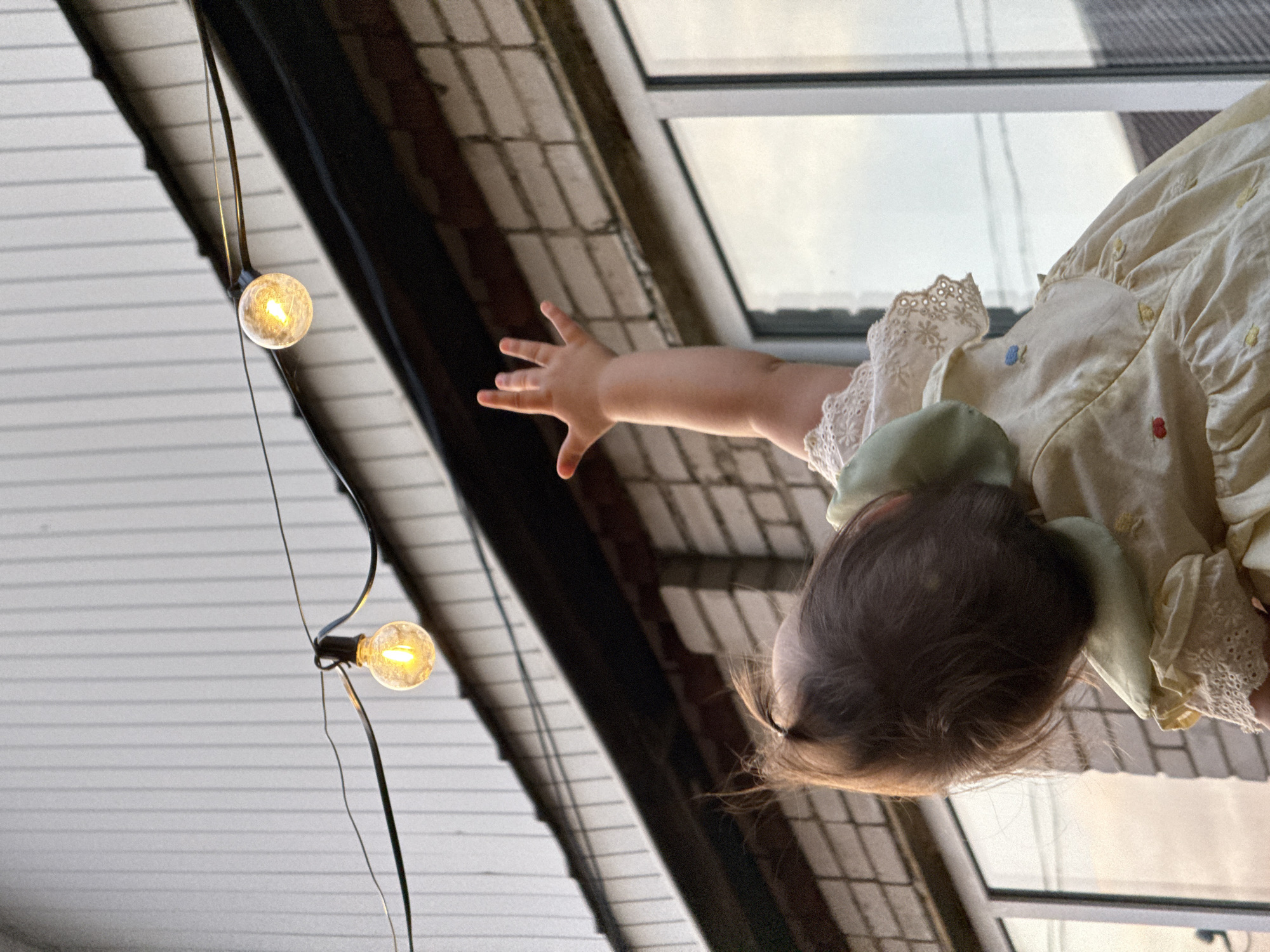 An overhead shot captures a young child in a light yellow dress reaching upwards towards two illuminated string lights. The child's back is to the camera, with their head tilted down and a single ponytail of dark hair visible. Their right arm is extended, with fingers spread. The dress features lace trim around the collar and sleeves, and small embroidered flowers in red and blue are visible on the bodice. Behind the child, a brick building with white trim and windows is visible, and above that, a white wall with vertical siding. The string lights are suspended on a black wire above the building. The scene is likely set outdoors, with the lighting suggesting it is dusk or evening. The image does not contain any specific landmarks or visual cues that would indicate its location within the city of Sharkan, Russia.