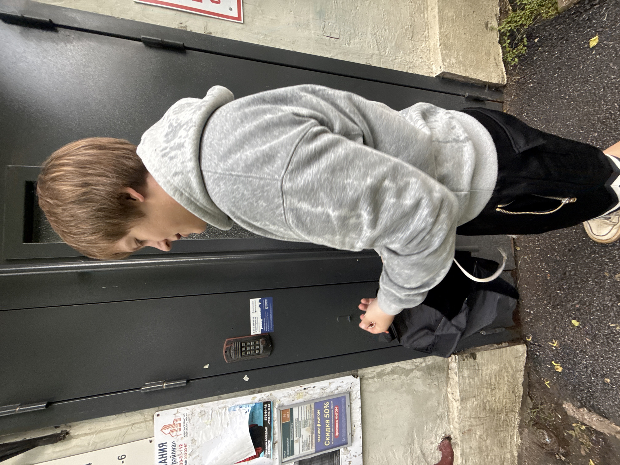 An individual stands on a wet asphalt surface in front of a dark gray metal door. The individual, seen from the chest down, is wearing a light gray hooded sweatshirt and black pants with a zipper detail on the side. They are holding a black bag, which appears to be an unfolded backpack, with their right hand. The door has a keypad entry system with red and black buttons mounted on it. To the left of the door, there are posters and flyers attached to a textured wall. The environment suggests an outdoor urban setting in Saint Petersburg, Russia, likely near an entrance to a building, with the ground wet from recent rain.