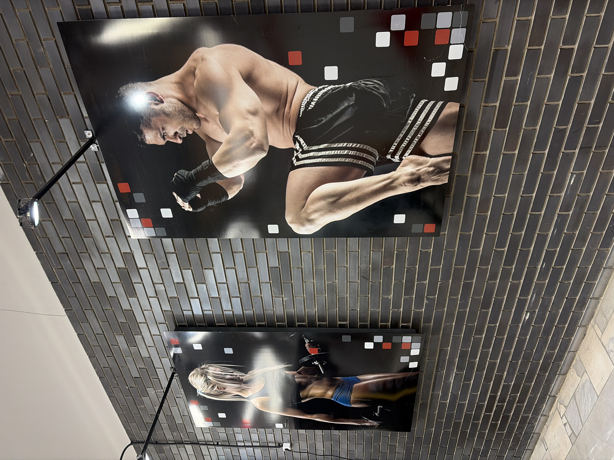 The image shows two framed posters on a tiled wall. The top poster features a shirtless male athlete in black shorts with white stripes, in a boxing or fighting stance. The lower poster shows a blonde woman lifting weights. Each poster has a pattern of red, white, and grey squares along the edges. A black adjustable lamp illuminates each poster. The tiled wall appears to be curved. The image was taken in Chaikovsky, Russia.