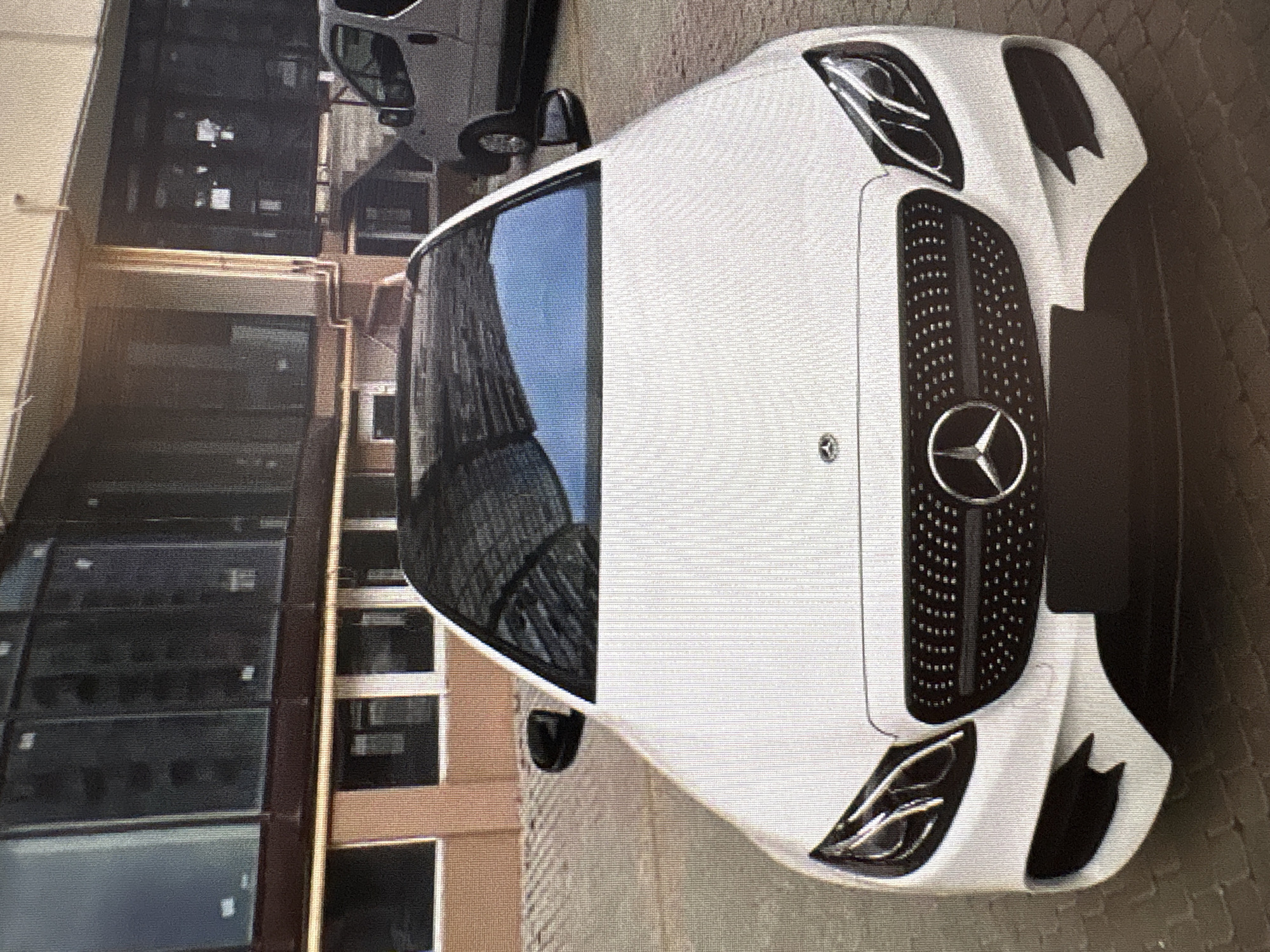 A white Mercedes-Benz E-Class sedan is parked on a brick-paved surface in front of a modern building with large windows. The car's front grille, headlights, and the Mercedes-Benz logo are visible. To the right of the car, a white panel van is parked. The building appears to be an office building or retail space. The scene is captured in daylight. The location is not identifiable as Moscow, Russia.