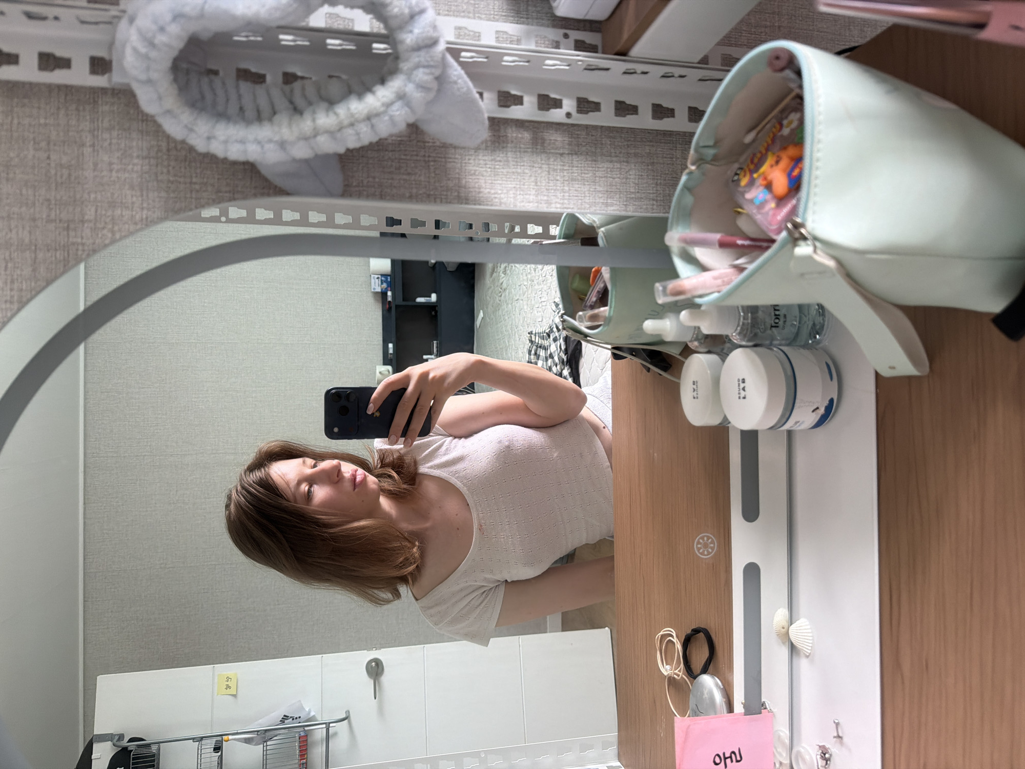 In a brightly lit room, a young woman with shoulder-length brown hair takes a selfie in a large, oval mirror. She is wearing a short-sleeved, white cropped top with a textured pattern. Her left hand holds a dark-colored smartphone. The mirror reflects her and the room behind her, revealing a white closet with a silver handle on the left. On the closet door, a small yellow sticky note is attached.
In the foreground, on a wooden surface, several items are laid out. A pale pink sticky note with Korean writing is visible, alongside hair ties and a silver compact. To the right, two white jars with blue and white labels sit next to a clear bottle with a blue label. A light teal makeup bag filled with cosmetics, including lipsticks and cotton swabs, is also present. The background shows a textured wall and a white shelving unit with a light blue fluffy headband hanging from it. The overall scene suggests a personal grooming area, likely in a residential setting in Dongducheon-si, South Korea.