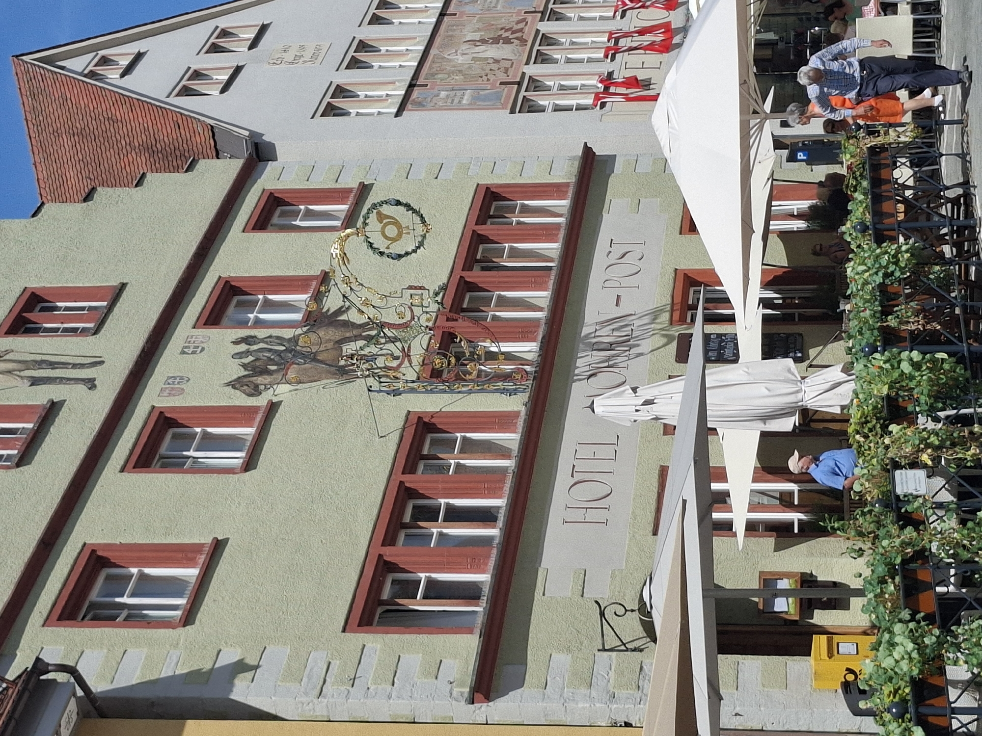 The image depicts the facade of the Hotel Mohren-Post in Wangen im Allgäu, Germany. The hotel is a multi-story building with a light green stucco exterior and red trim around the windows. A historical mural depicting a postilion on horseback is visible on the upper part of the building, alongside a decorative post horn sign. The ground floor features a restaurant or cafe area with tables and umbrellas. A man in a blue shirt and a cap is standing near a yellow mailbox. Further to the right, an older man in a checkered shirt and dark pants walks past tables. Several people are seated at tables, and red and white flags are hanging from an adjacent building. The street is paved, and lush green plants line the foreground. The sky is clear and blue.