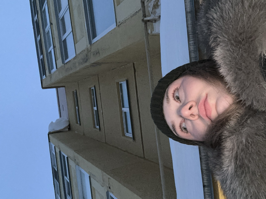 A woman wearing a dark knitted hat and a fur-lined coat poses in front of a multi-story building with many windows. The building's exterior is a beige color. The sky is visible behind the building. A patch of snow is on the building's roof. The woman's face is tilted towards the camera, and her expression is neutral. The image is taken from a low angle, making the building appear taller.