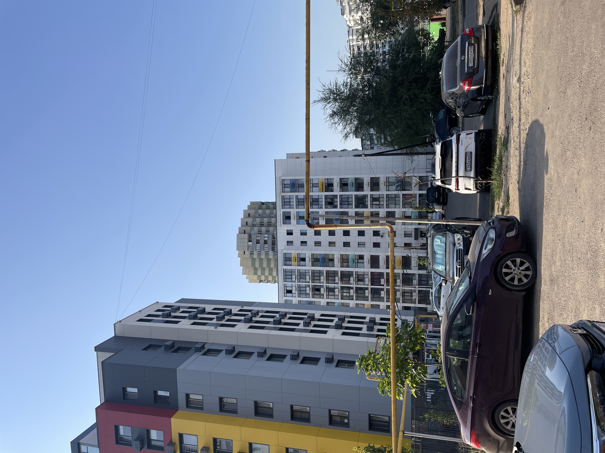 The image captures a street scene in Almaty, Kazakhstan, on a clear, sunny day. In the foreground, a dark purple sedan is parked on a dirt lot, with the front of a grey car visible to its left. Behind these cars, several other vehicles are parked in a line, including a white SUV and a dark sedan. The backdrop is dominated by modern apartment buildings with distinct architectural features. The building on the left is a multi-colored structure with sections of yellow, grey, and red. Adjacent to it is a white building with numerous windows and balconies. Further in the distance, another tall building with a patterned facade is visible, along with more apartment blocks. A yellow pipe runs horizontally across the middle of the frame, supported by posts, in front of the buildings. Green trees and foliage are interspersed between the buildings and the parking area. The sky is a clear, bright blue. There are no people clearly visible in the scene.