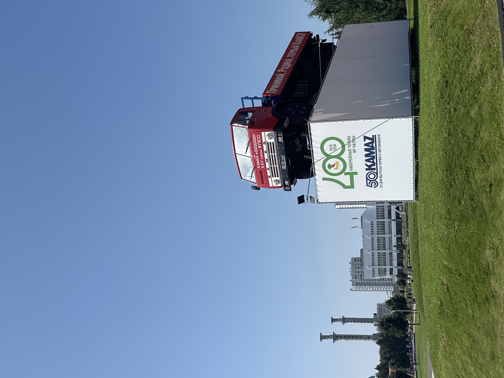 The image shows a red and white Kamaz truck mounted on top of a large white banner display in Naberezhnye Chelny, Russia. The banner features the Kamaz logo, the text "50 KAMAZ" with "СО ДНЯ ВЫПУСКА ПЕРВОГО АВТОМОБИЛЯ" below it, and another logo with the text "1626 2026 НАБЕРЕЖНЫЕ ЧЕЛНЫ ЯР ЧАЛЛЫ". The truck has the text "КАМАЗ № 0000001" and "XXV съезд КПСС" on its windshield, and "00-01 TAT" on its license plate. The banner also has "ПРИНИМАЙ, РОДИНА, ПЕРВЫЙ КАМАЗ!" written in red on the side of the truck. The truck and banner are displayed outdoors on a sunny day with a clear blue sky. In the background, there are several buildings, including a modern white building and two industrial-looking towers. A grassy lawn is in the foreground. There are a few individuals walking on the lawn in the middle ground.