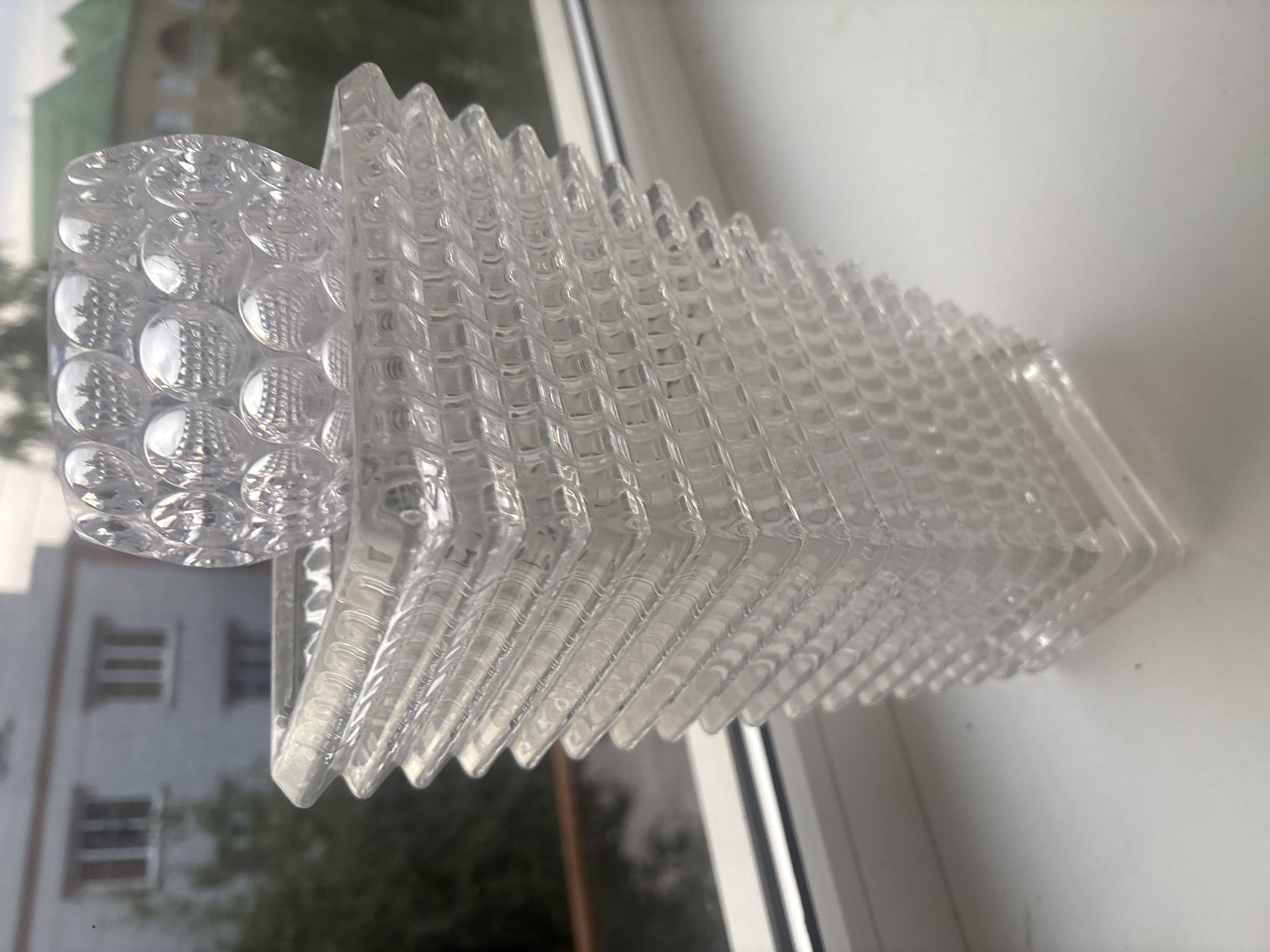 A clear glass vase with a patterned texture sits on a windowsill. The vase is square at the base and tapers upwards to a wider opening, with a layered, stepped design. A circular, bubble-patterned glass ornament rests on top of the vase. In the background, through the window, a building with multiple windows and a green roof is visible, along with trees and foliage. The scene suggests an indoor setting looking out onto an urban environment, likely in Georgiyevsk, Russia, given the context. There are no people present.