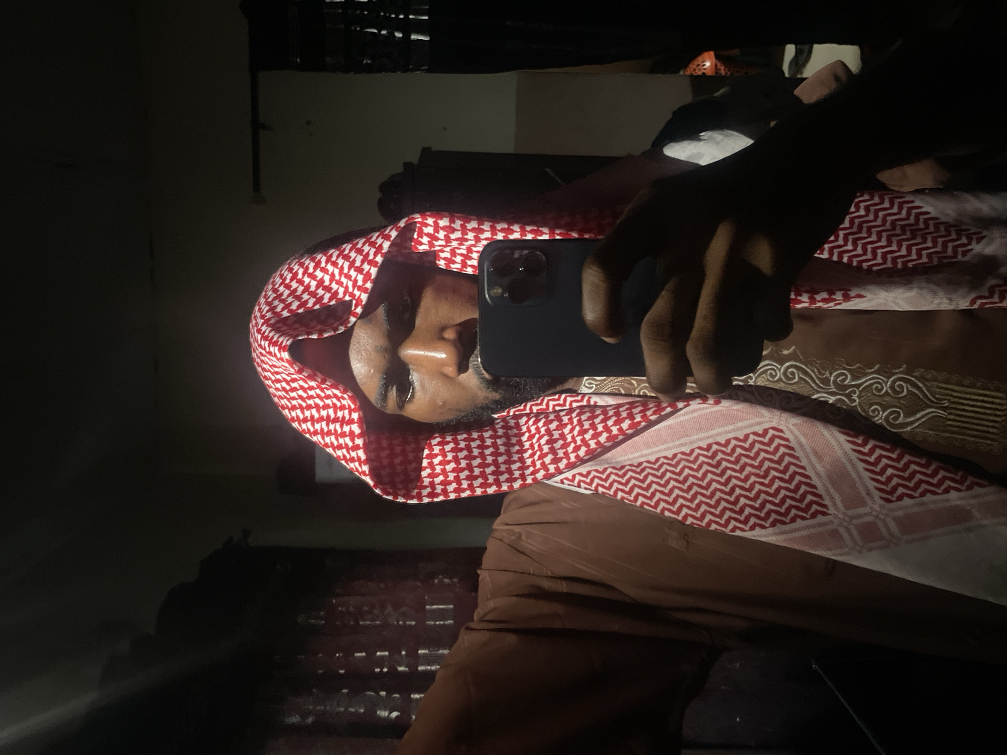 A person in a red and white checkered keffiyeh is taking a selfie with a dark-colored smartphone. The person is wearing a brown garment with decorative patterns. The background is dark and indistinct, but a blurred structure with horizontal bars is visible. There is no specific information available in the image to confirm it was taken in Lokoja, Nigeria.