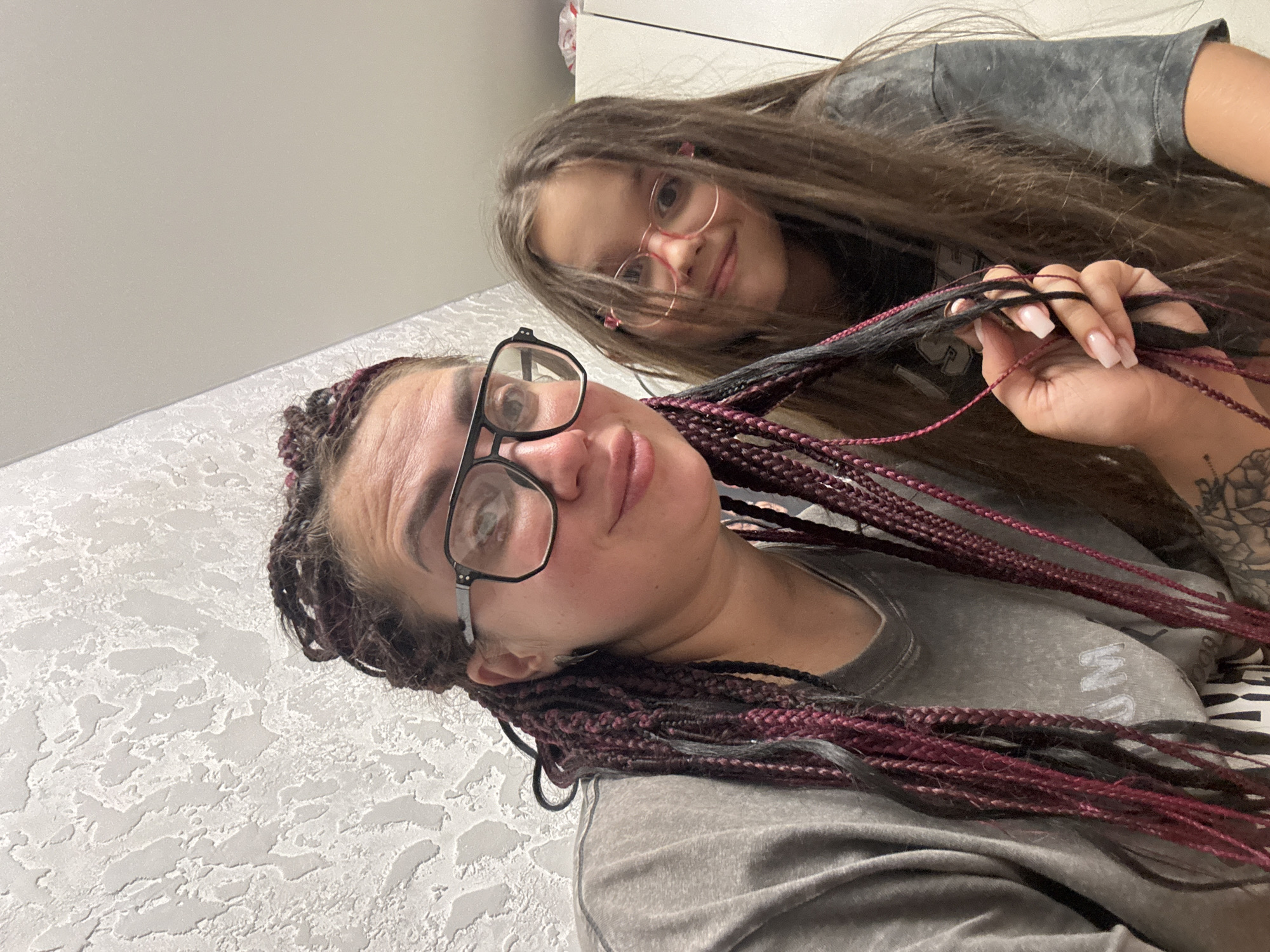 This is a close-up, selfie-style photo of two females, likely a mother and daughter, indoors. The woman in the foreground, on the left, has dark hair styled in numerous braids with reddish-purple highlights. She is wearing glasses with thick, black frames and a gray t-shirt. Her right hand is visible, touching her hair. The girl on the right has long, brown hair and is wearing pink-rimmed glasses and a gray t-shirt. She is smiling and looking towards the camera. The background consists of a textured, light-colored wall and a hint of white cabinetry. There is no identifiable information about the city of Tolyatti, Russia, within the image.