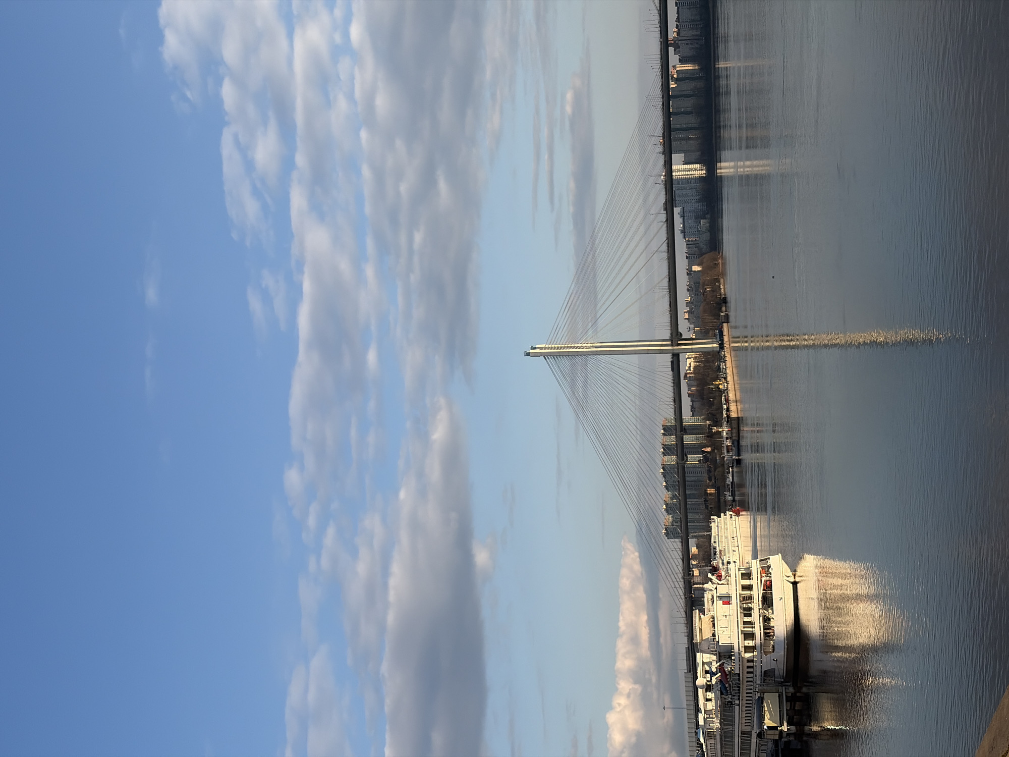 The image shows a view of the Neva River in Saint Petersburg, Russia, with a cable-stayed bridge in the background. The bridge appears to be the Cable-stayed Bridge, or Kantemirovsky Bridge. In the foreground is a large, white, multi-deck passenger ship docked at a pier. The river reflects the bridge and buildings on the opposite bank, as well as the cloudy sky above. The sky is blue with scattered white and gray clouds. The orientation of the image is vertical.