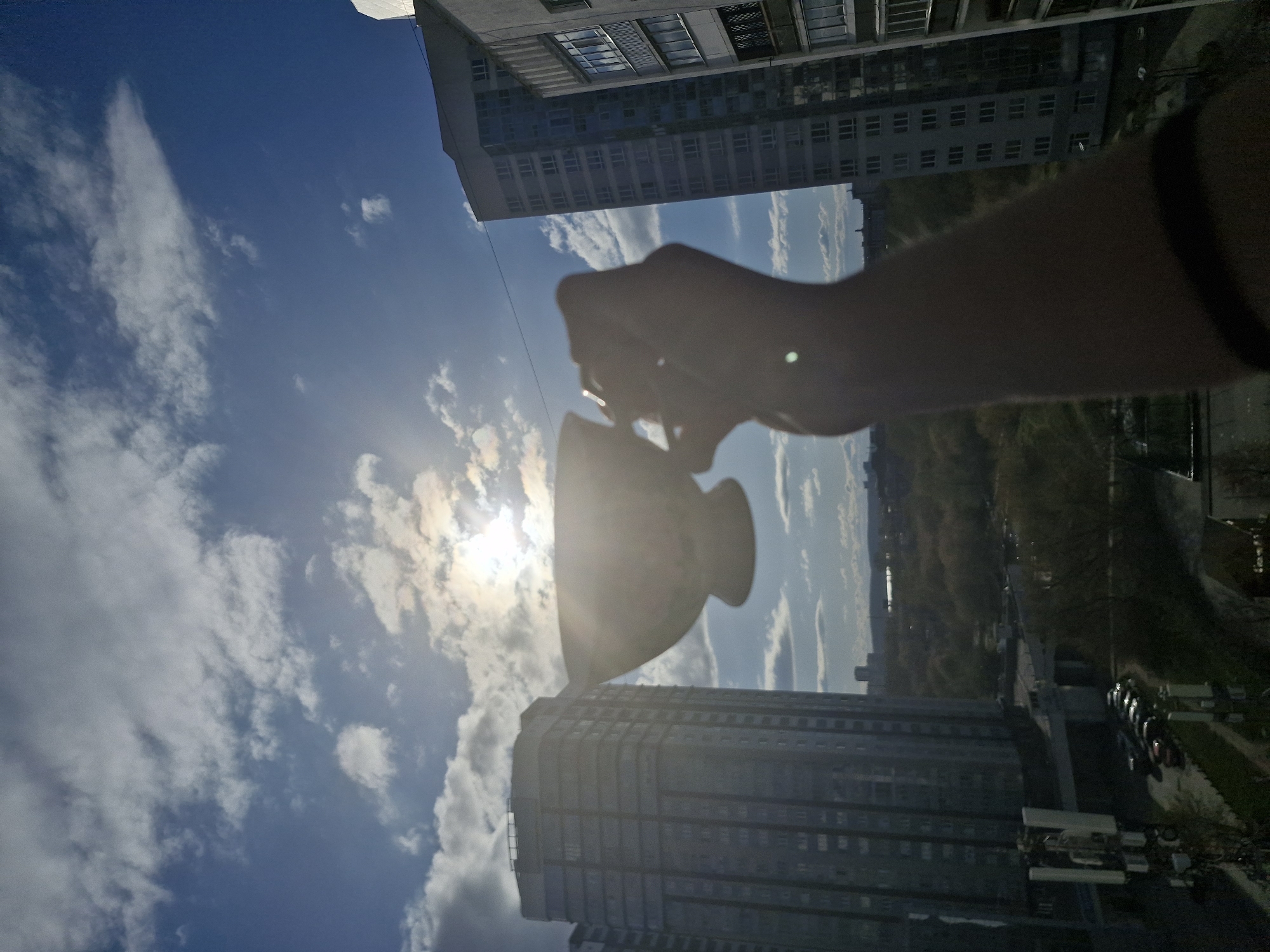 This image captures an outdoor urban scene, presented rotated 90 degrees clockwise. In the foreground, a person's arm and hand are prominently silhouetted against a bright sky. The hand holds a small, decorative object, possibly a vase or urn, which is also silhouetted. A dark wristband is visible on the person's wrist.
The sky is a clear blue with scattered white and grey clouds. The sun is intensely bright and positioned behind the held object, causing significant lens flare and creating a strong backlighting effect on the foreground elements.
Below the sky and extending into the background are several urban buildings. A tall, modern high-rise building with numerous windows occupies the left side of the frame. Another multi-story building, possibly residential or commercial, is visible further back and to the right. Beyond the buildings, a faint landscape suggests distant trees or urban structures, and possibly a road or body of water.
The scene depicts a deliberate photographic act, where the object is held up to the sun to create an artistic silhouette. The time of day is bright daytime, characterized by strong sunlight and a partly cloudy sky. The setting is an urban environment, with the provided context placing it in Yekaterinburg, Russia. There is no discernible text visible in the image.