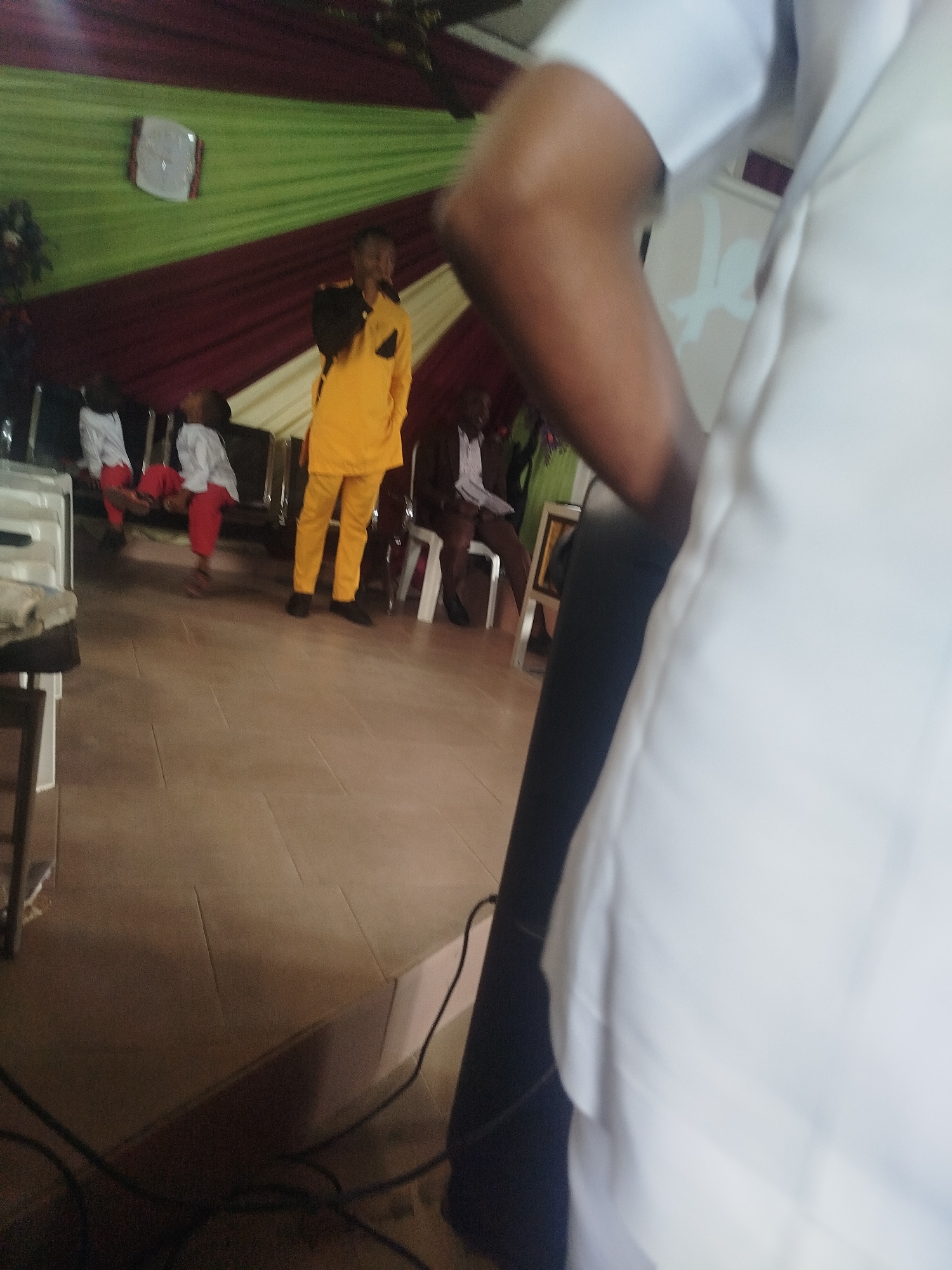 The image depicts a scene in a room, possibly a church or event space, located in an unknown city. The background features a curtain with green, maroon, and white stripes. A man in a bright yellow suit stands on a slightly elevated platform, possibly speaking or performing. Two children dressed in white shirts and red pants are visible to the left of the man, engaging in some activity. Another man is seated on a chair in the background, wearing a suit and tie. The foreground is dominated by a large white object, possibly a piece of clothing, obscuring part of the scene. A black cable runs along the floor. The setting appears to be indoors with tiled flooring.