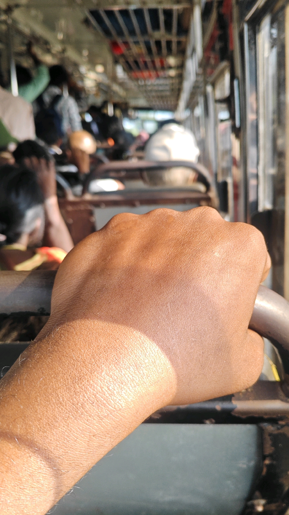 The image shows a close-up of a person's hand gripping a metal bar inside a crowded bus. The hand is tanned and shows visible skin texture and some hair. Sunlight casts a shadow across the hand. In the background, the interior of the bus is visible, with other passengers and their belongings, suggesting a busy journey. The setting appears to be during daylight hours, with a glimpse of the outside through the bus windows. The overall impression is one of a common travel scene on a public transport vehicle in India, likely within the city of Virat Nagar, given the context.